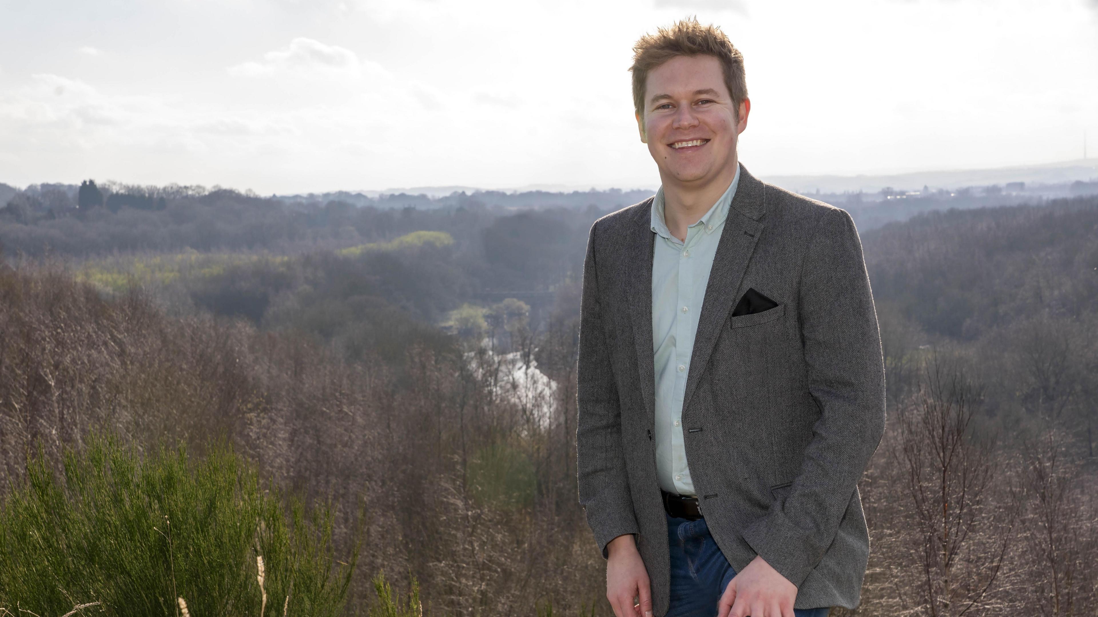 Councillor Jack Hemingway, a tall young man in a grey suit jacket, pictured against a large clump of trees on the slopes and hills behind him. He is smiling into the camera.