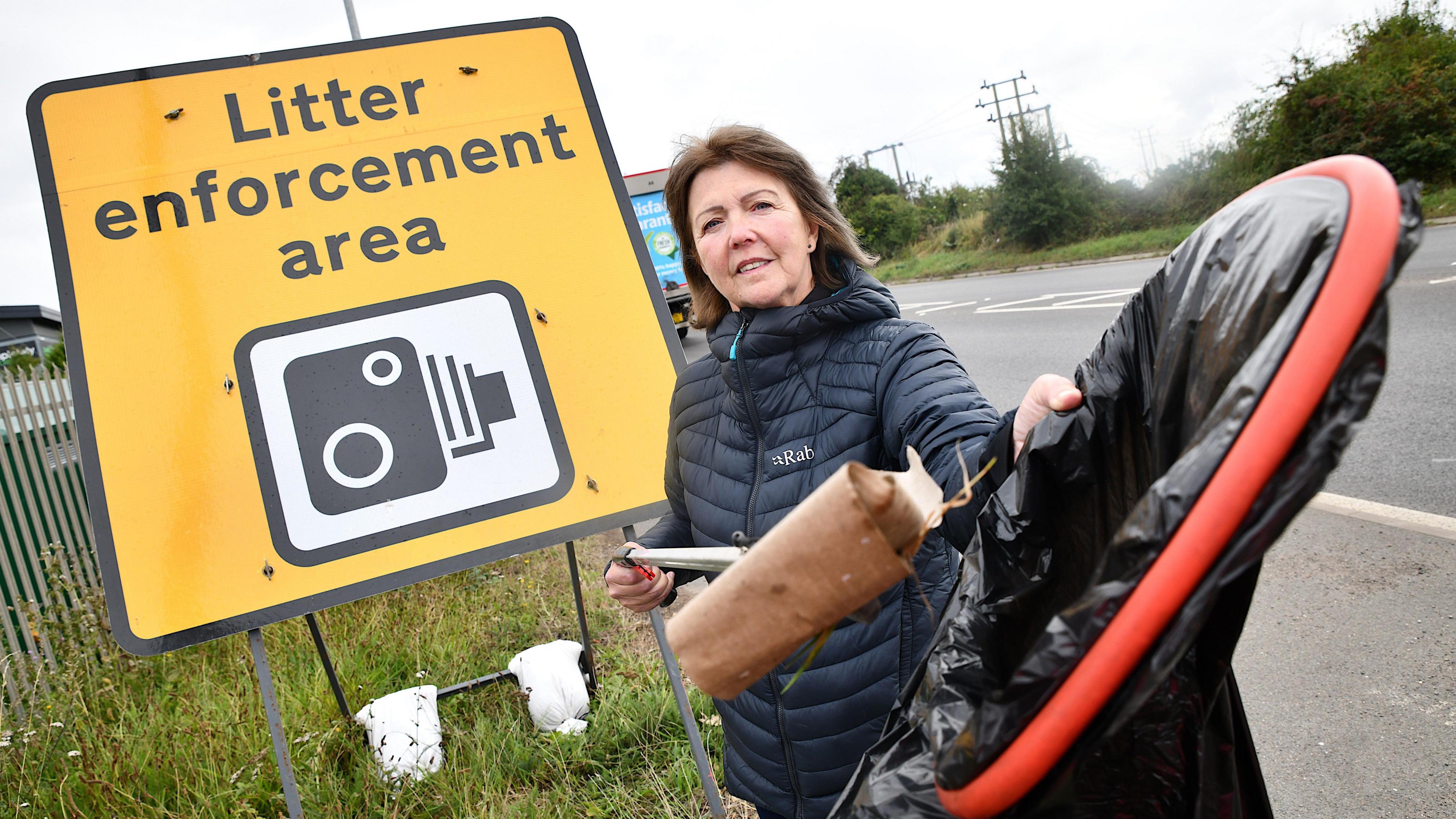 Lynda Hodgkins with a litter picker standing in front of the traffic enforcement area sign on the A5