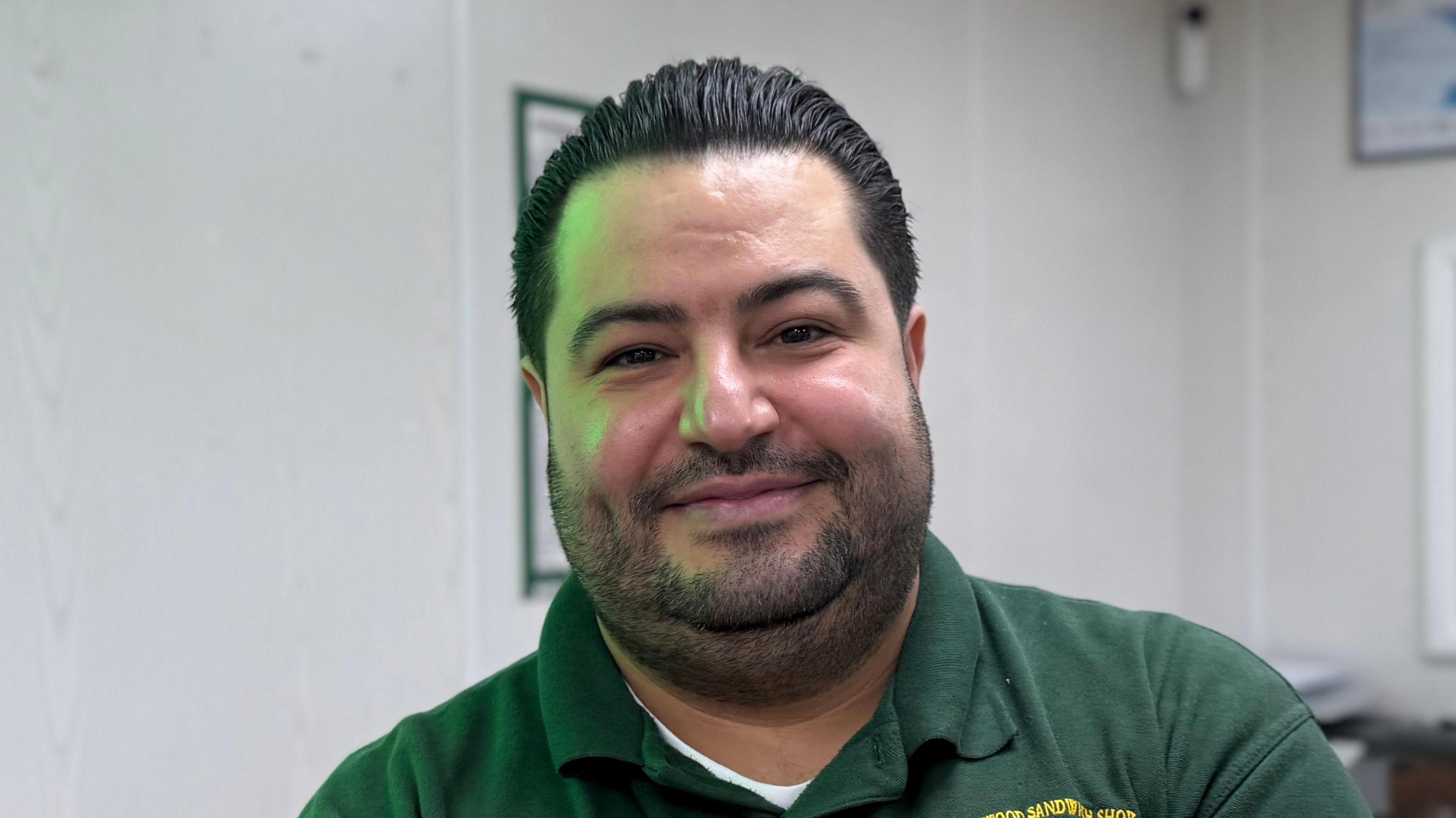 A man - Murat Mutlu - looking straight at the camera with a happy expression. He is standing in a sandwich shop and is wearing a green polo shirt with the name of the shop - Honywood Sandwich Shop - embroidered in yellow.