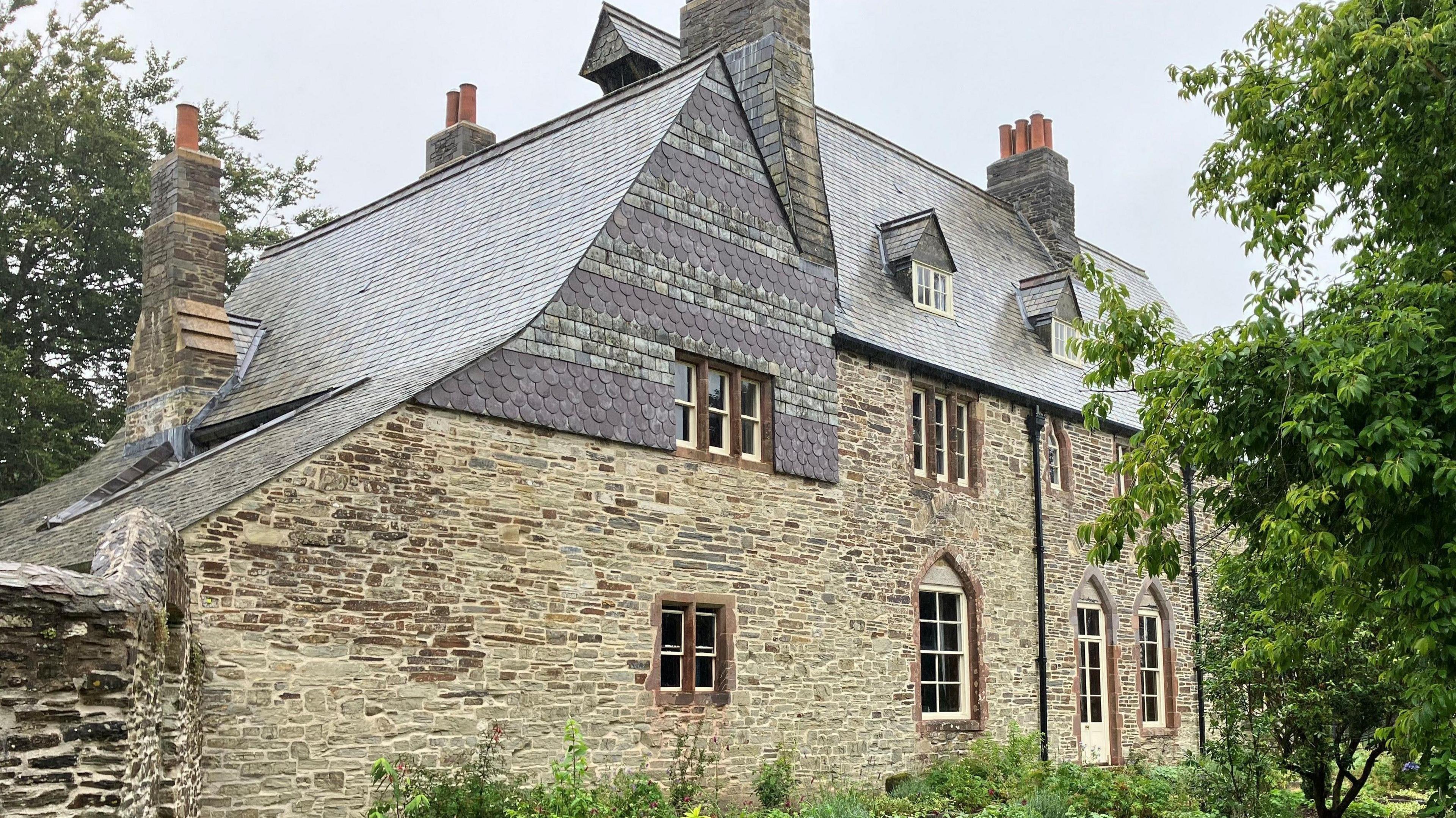 A house made of stone with a slate roof and three chimneys and multiple windows. There is a tree in the foreground.