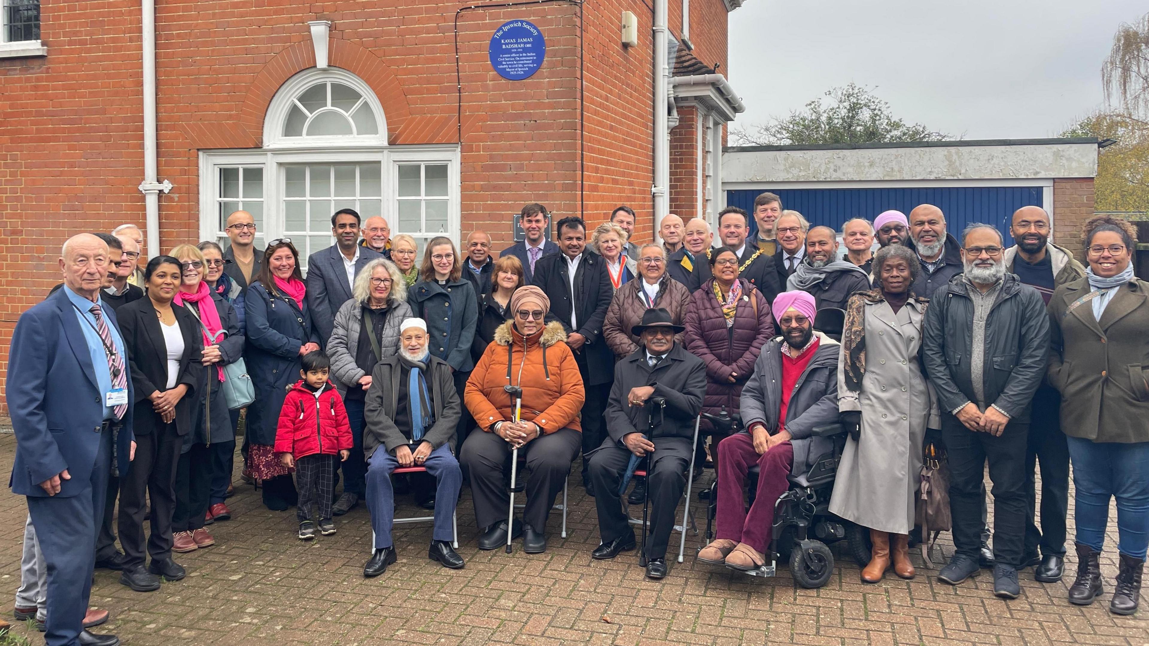 A group of about 50 people bunched together outside, smiling at the camera. Behind them is a red brick house with a blue circular plaque on the wall.