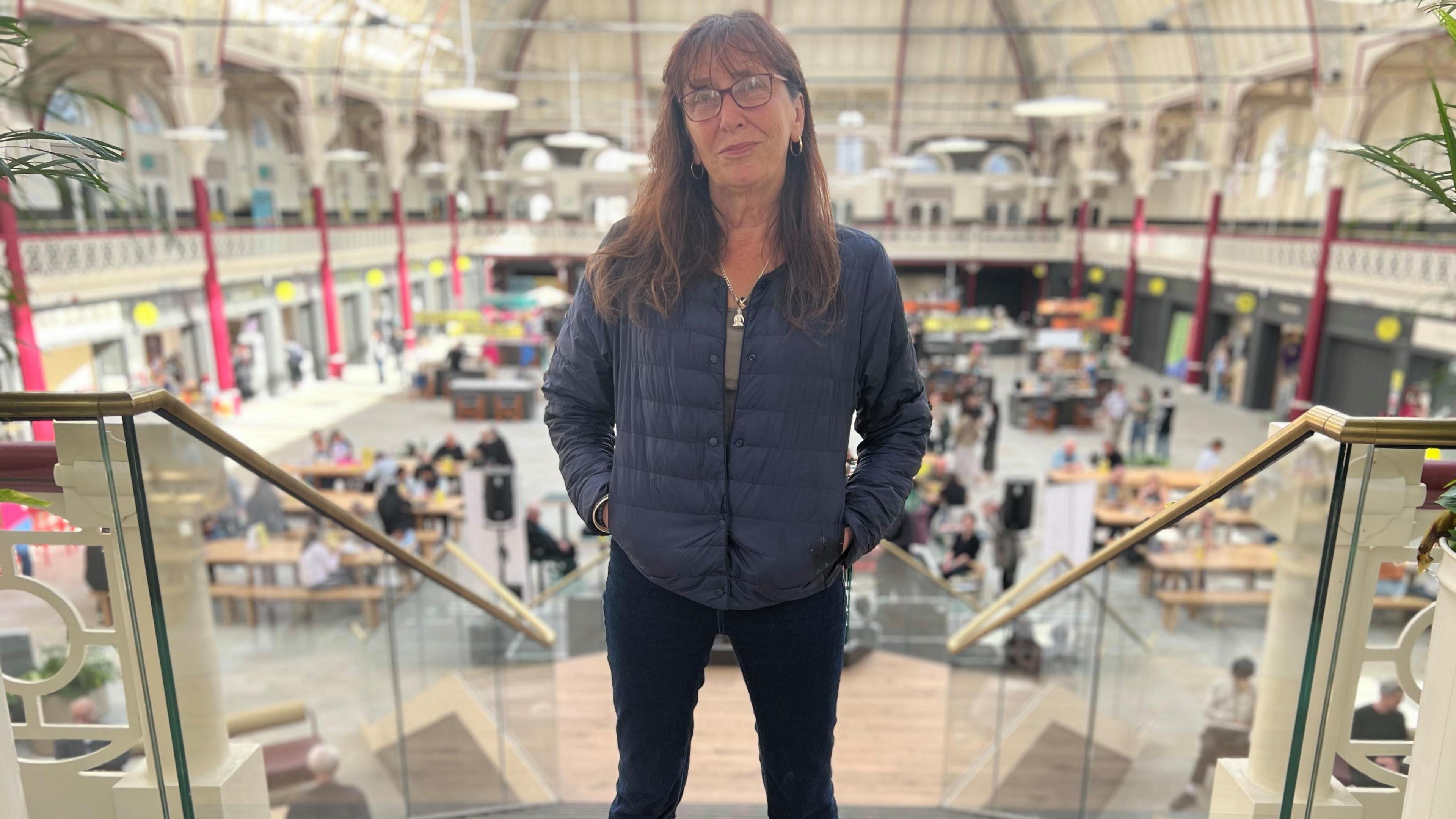 Woman with long brown hair and glasses in blue puffer jacket and jeans stood on staircase with view of Market Hall floor behind her