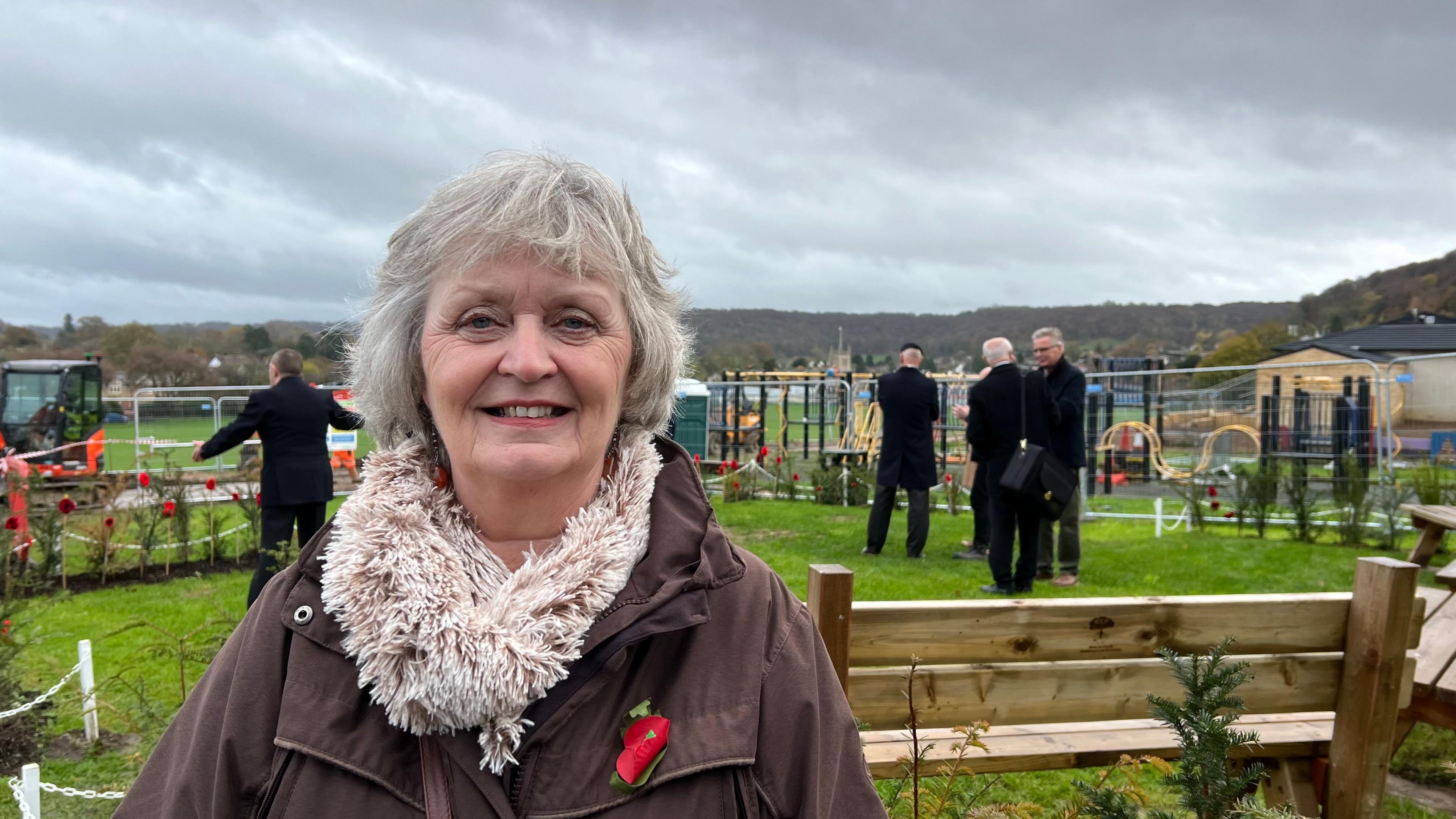 A women is stood in front of the new war memorial garden in Dursley. She is looking at the camera and smiling. She is wearing a poppy on the left collar of her brown coat and beige scarf. 