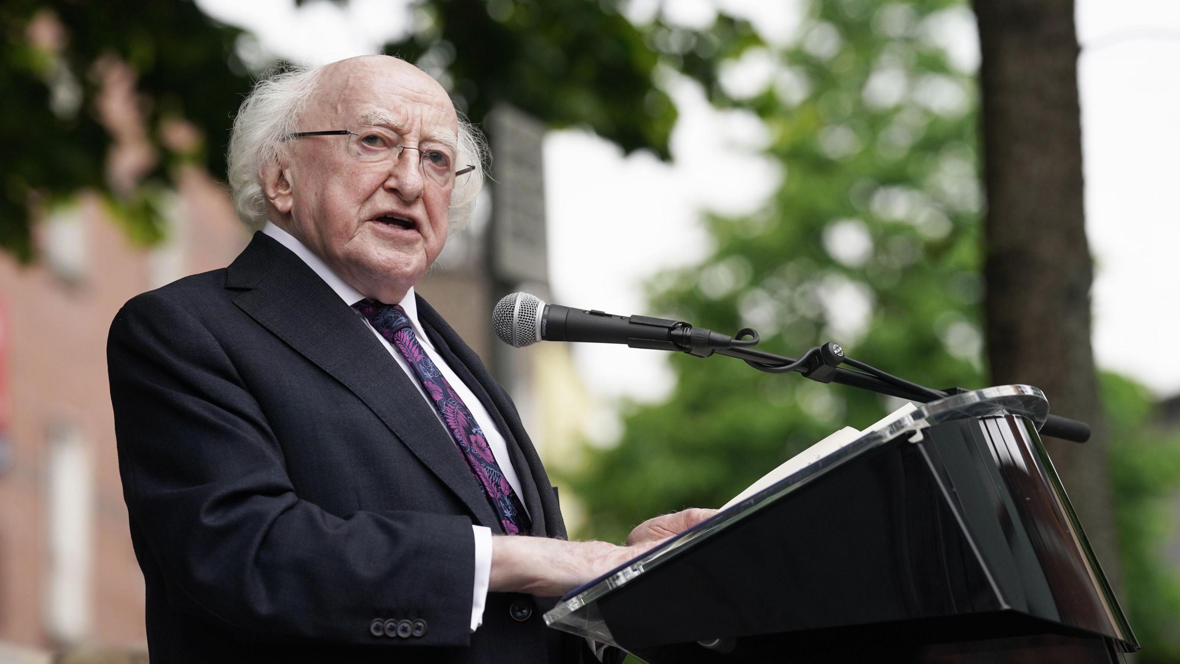 President of Ireland Michael D Higgins speaks at a lectern. We wears a black suit with a white shirt and and a purple floral tie. 