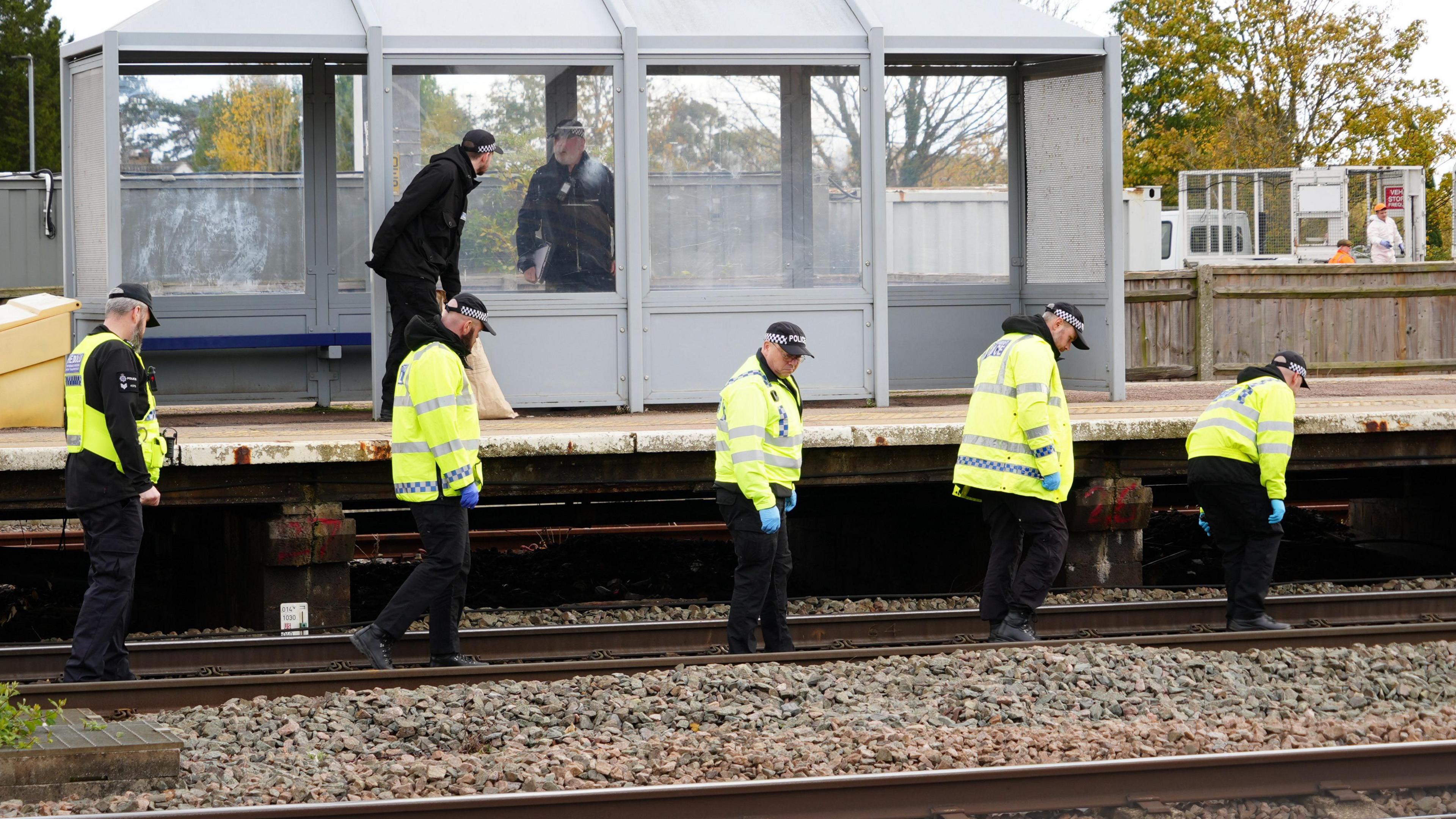 Five police officers walk along the track wearing high vis coats and black police hats. They are all staring down at the track looking for clues. The platform is next to them, with a plastic shelter, where two other officers are talking.