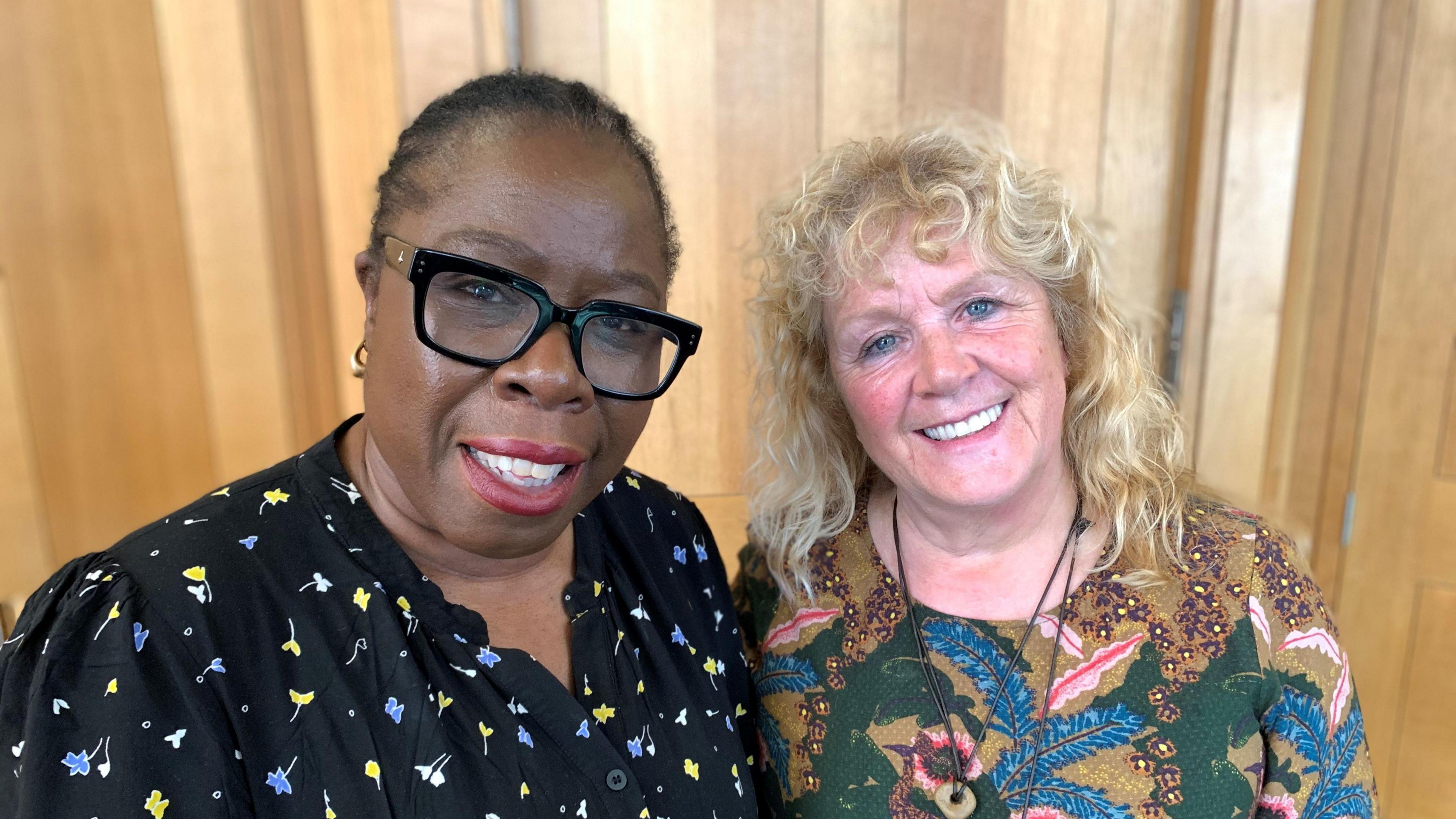 Two women stand next to each other, smiling at the camera. The woman on the left wears a black shirt with yellow and blue flowers on it, and black glasses. The woman on the right wears a colourful patterned shirt.