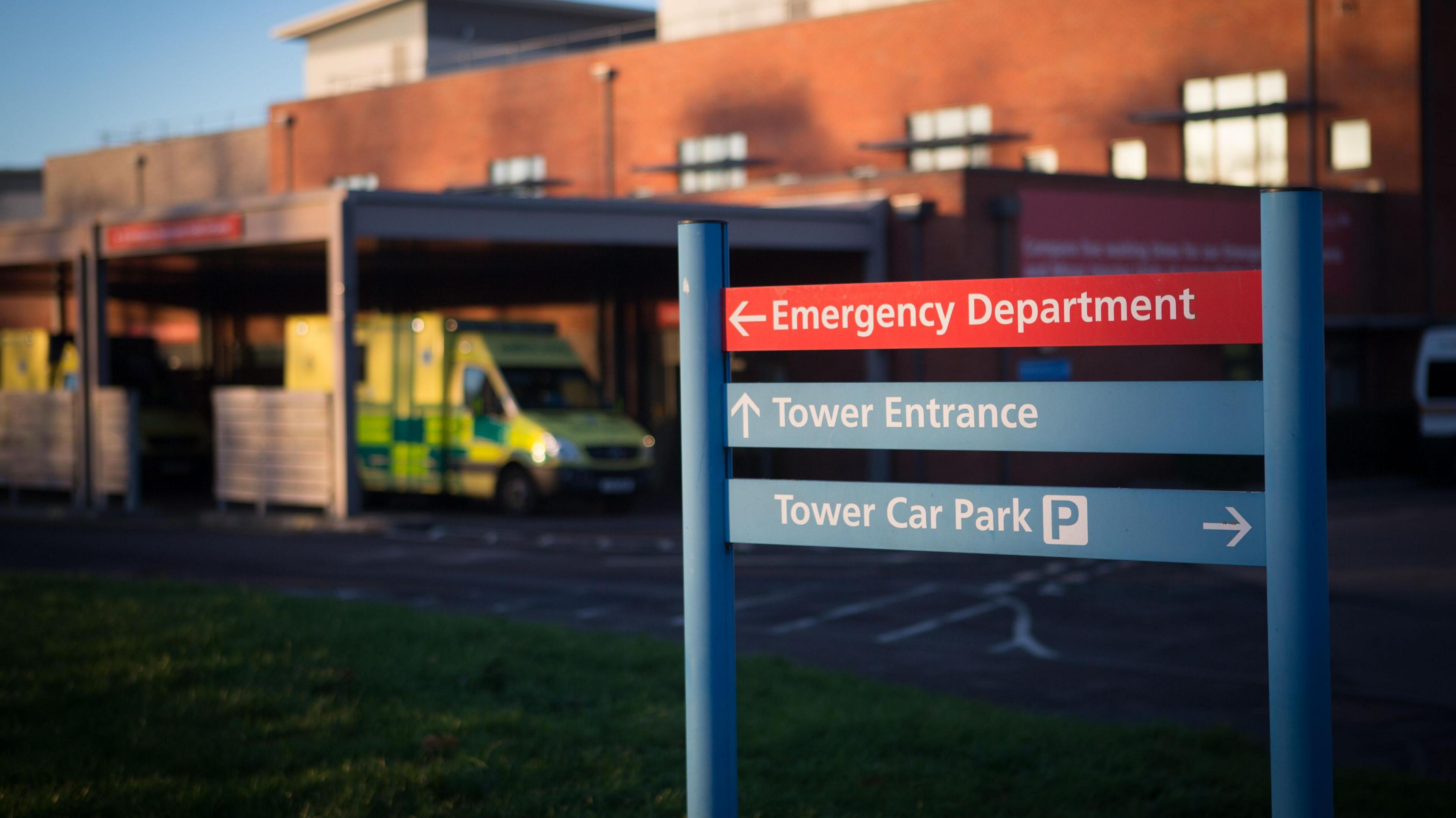 A sign in front of a hospital, with an ambulance parked in front of it.