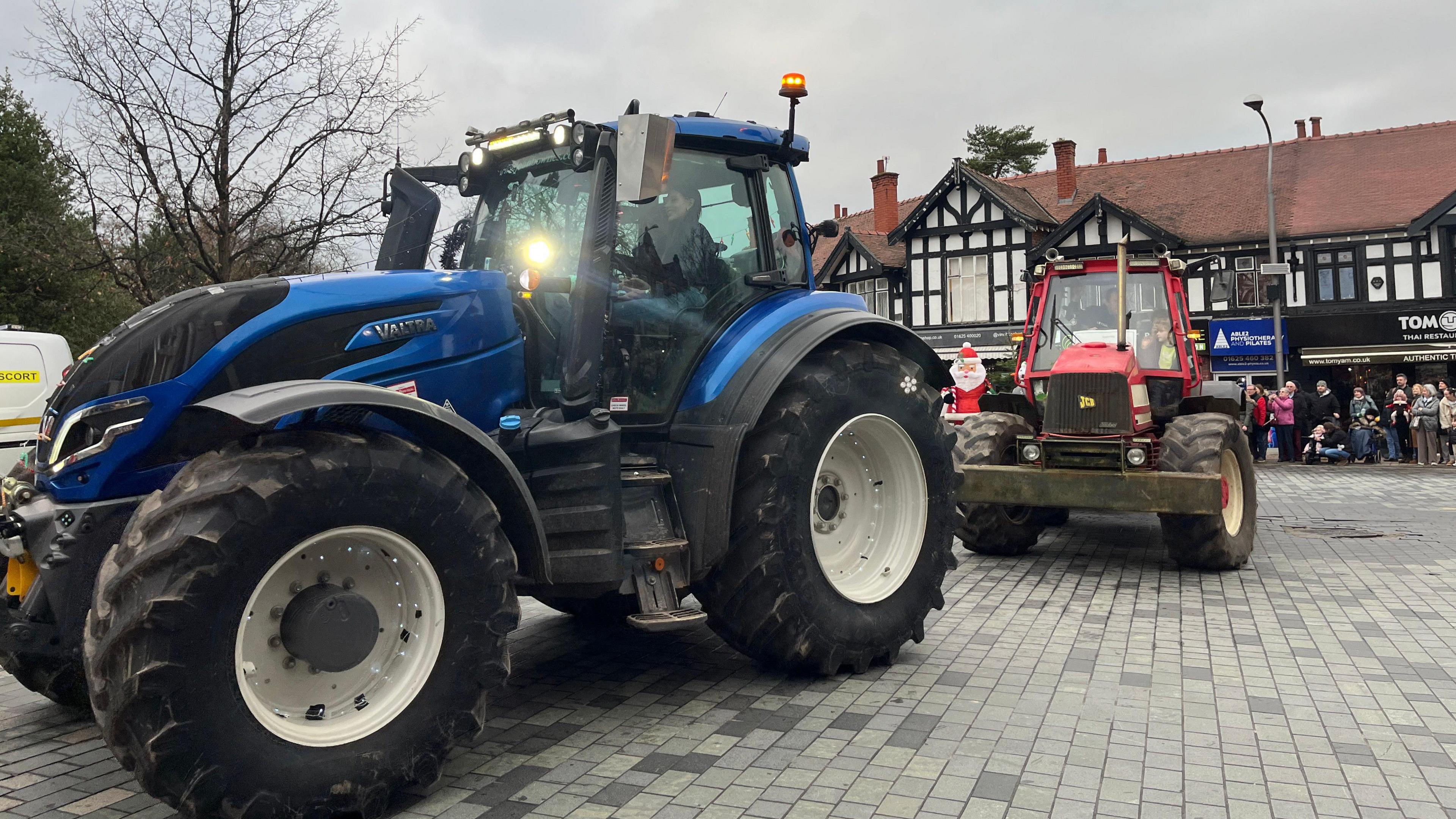 A giant blue tractor sits in front of a red tractor in a town square, with crowds looking on 