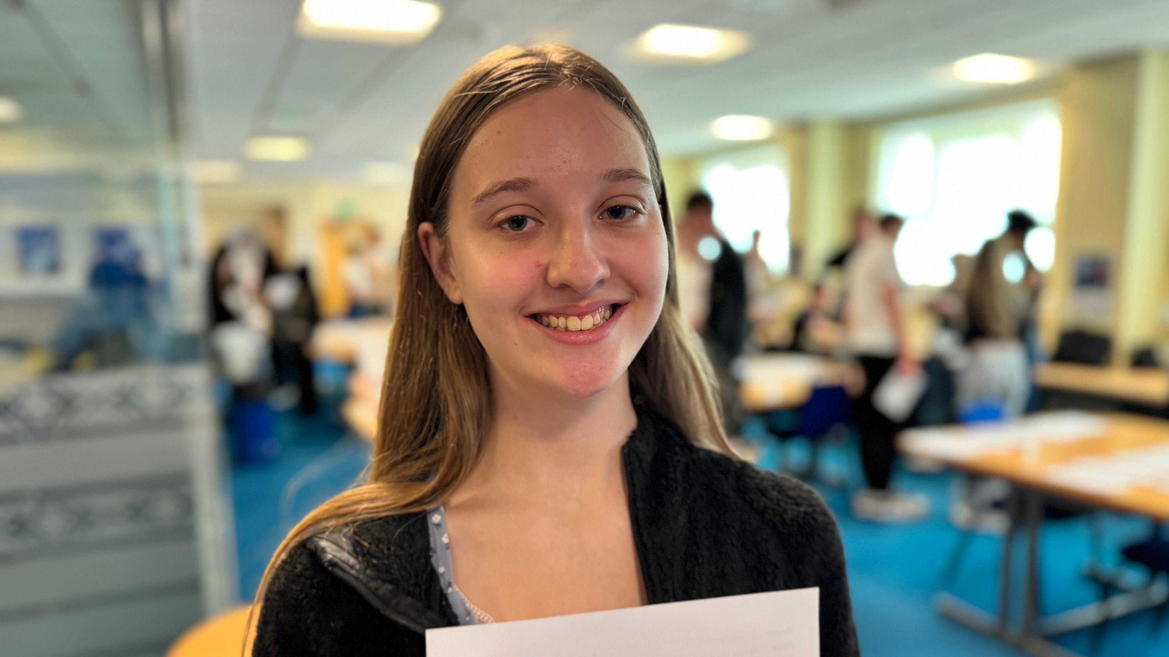 Jennifer Mordue smiles and holds her results on a white piece of paper. She has long brown hair and wears a black cardigan. She stands in a sixth form college while other pupils look for their results in a blurred background.