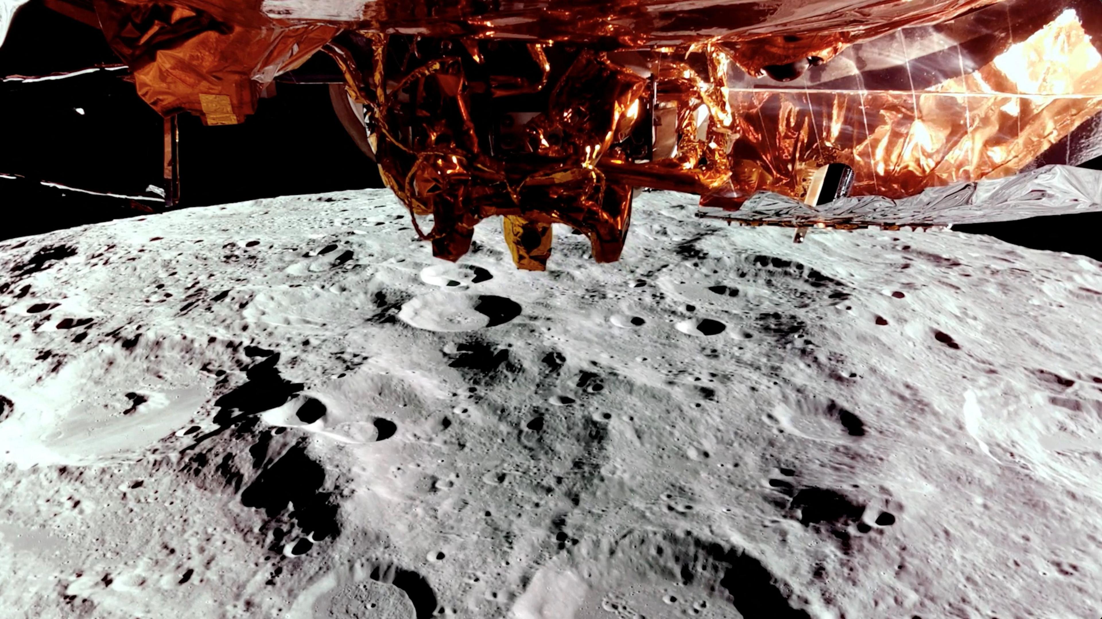 The Moon passes underneath the Blue Ghost spacecraft on 24 February 2025 as it orbits the lunar surface ahead of its landing on 2 March 2025. The gold foil underneath the spacecraft is visible, below is the cratered gray surface of the moon and the blackness of space is in the background. 