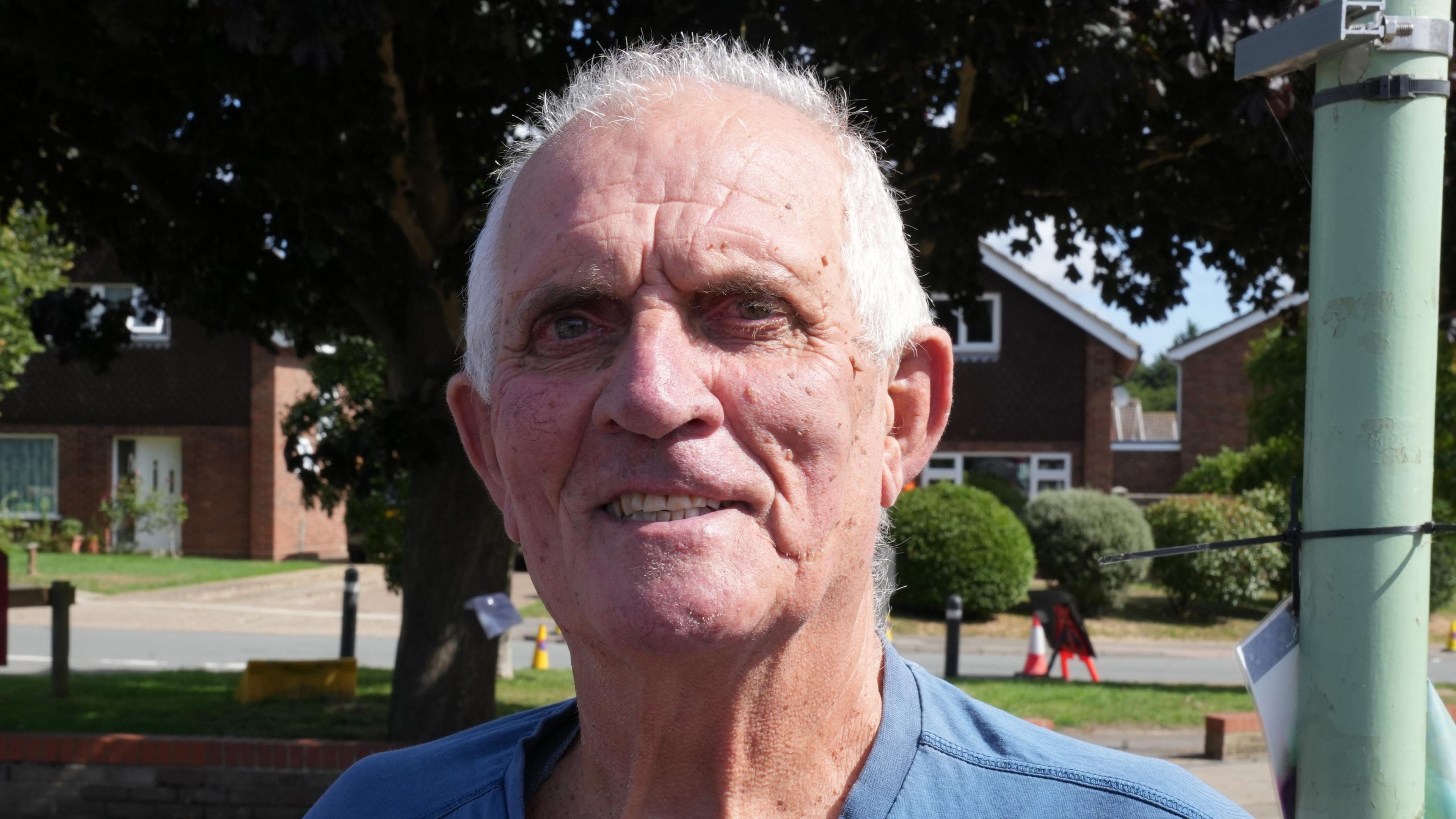 A man smiles at the camera. He has short, thin grey hair and is wearing a blue T-shirt.