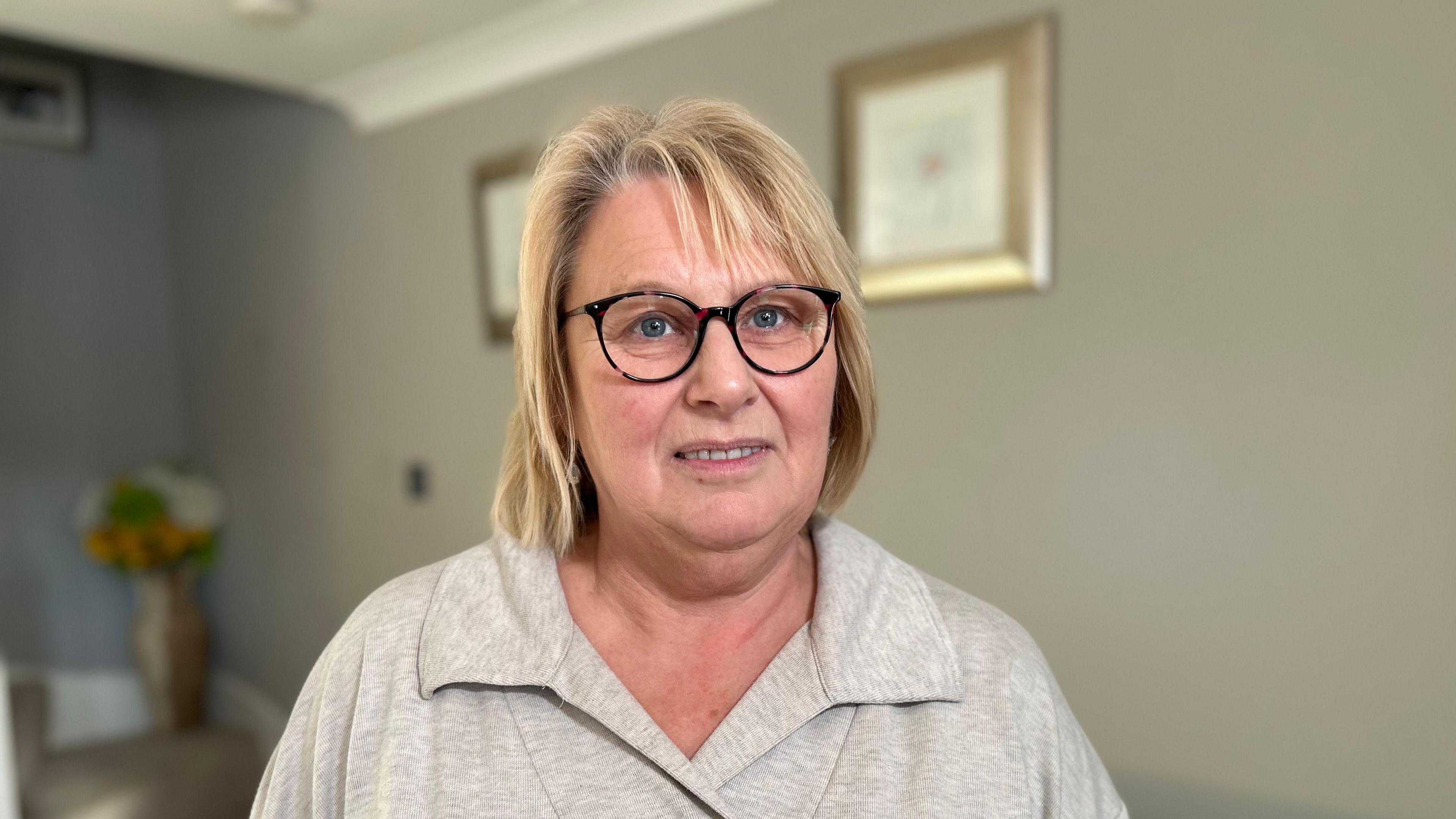 A woman with shoulder length blonde hair smiles at the camera. She is standing in a living room and is wearing black glasses with a grey top on.