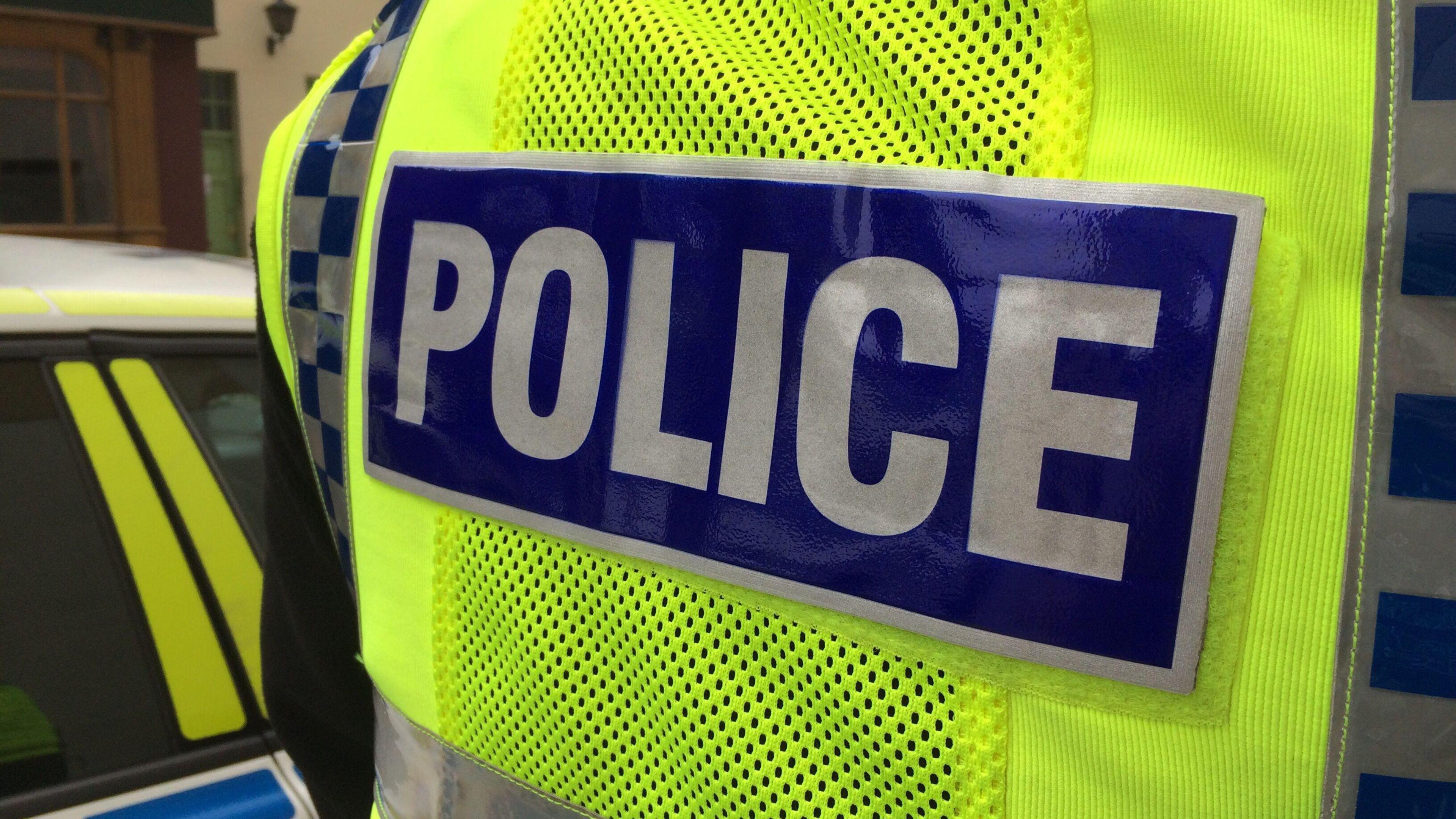 The back of a police officer's high-visibility yellow jacket. The words POLCIE are written in block capitals, stitched on to the back of the jacket.