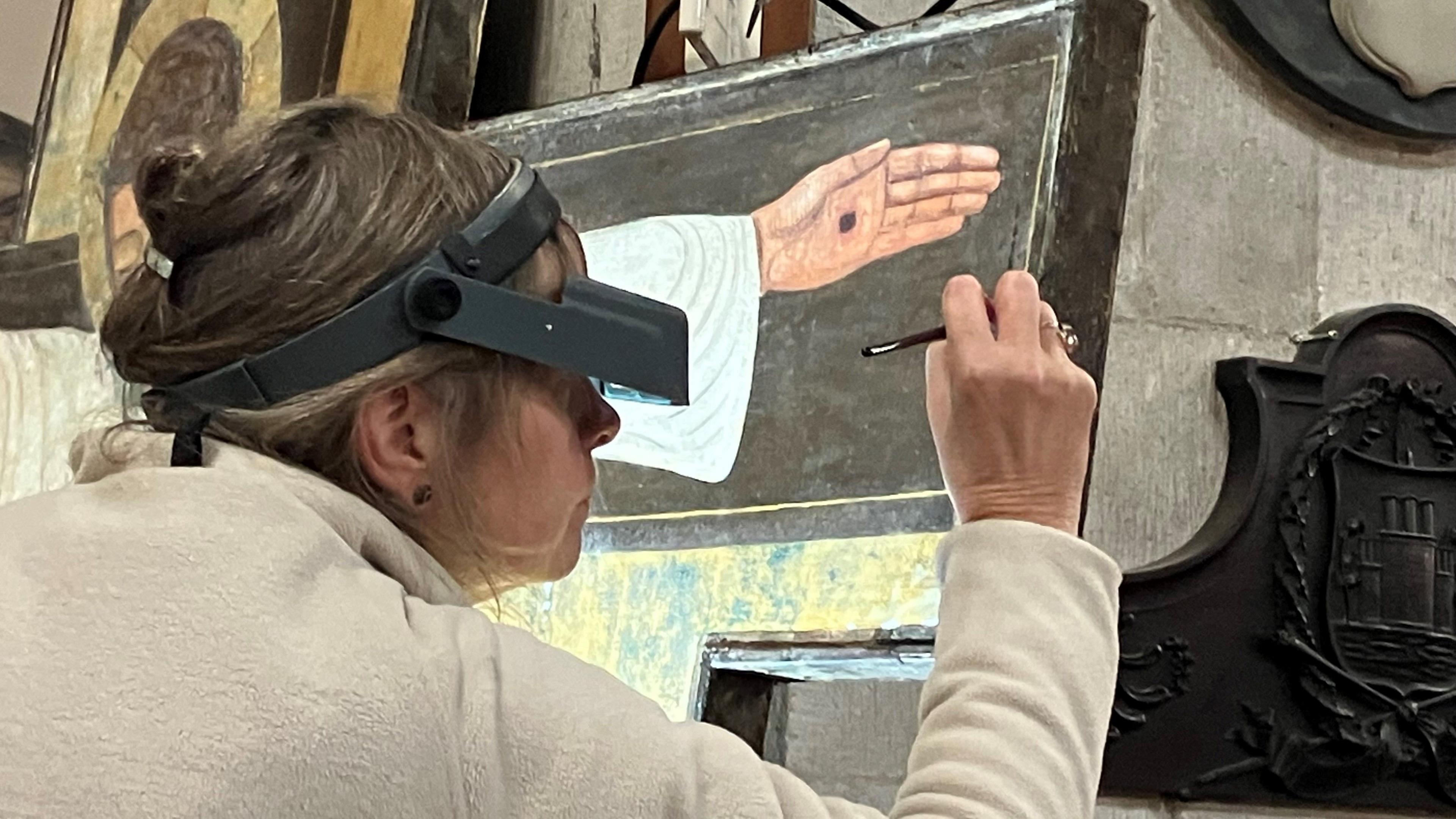 Woman with her hair tied up and wearing a magnifier on her head in order to see a close up of the painting she is restoring. She has a paintbrush in her hands and is working on the side of the painting. You can see part of the painting which depicts Jesus Christ's hand and a black dot which represents the crucifixion