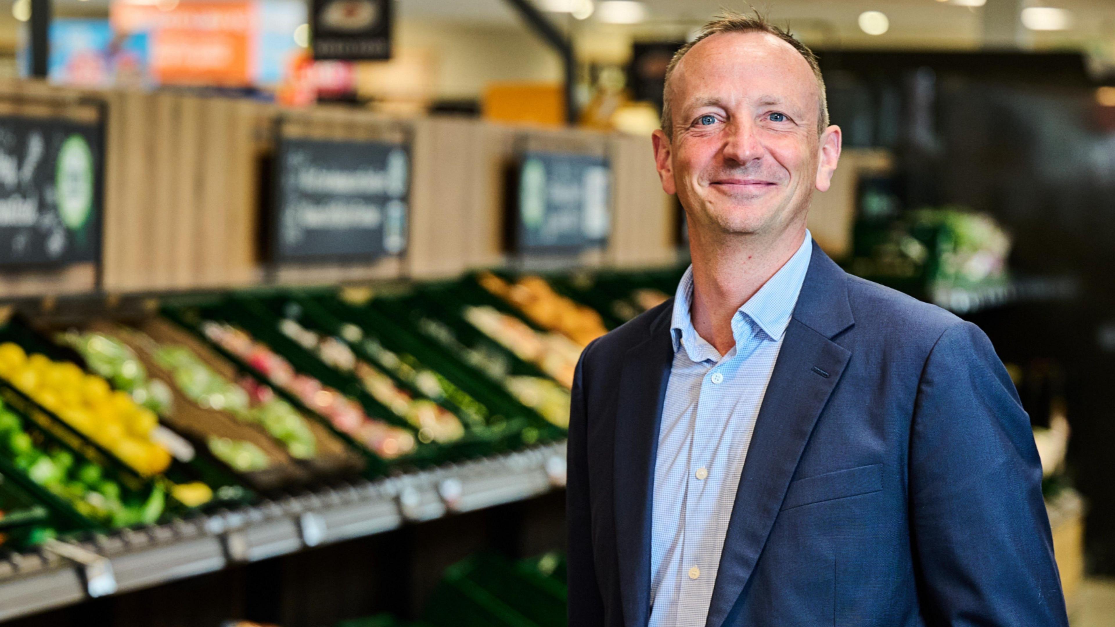 The chief executive of Aldi UK, Giles Hurley, smiles for a photograph in the vegetable aisle of an Aldi supermarket while wearing a blue shirt with no tie and blue blazer