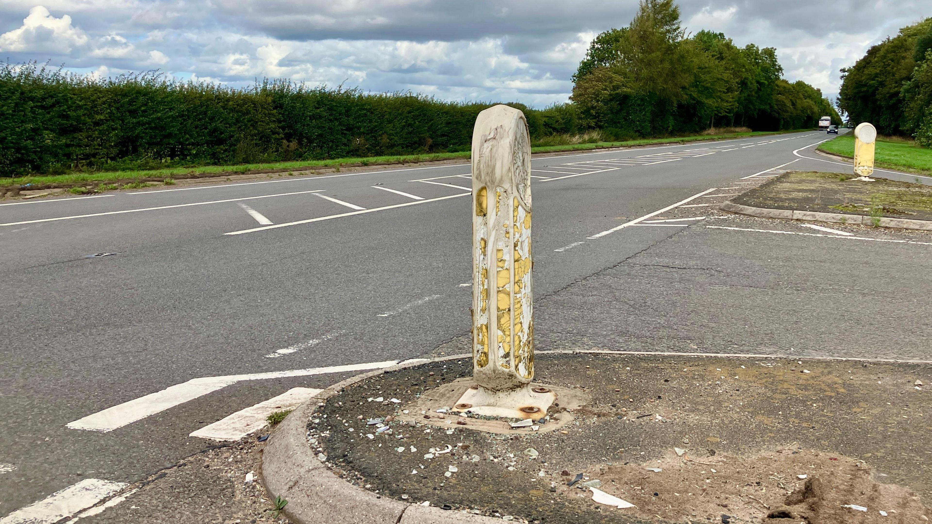 A shabby yellow and white bollard, with shattered pieces around it, at a T-junction, with bushes and trees on the other side of the road