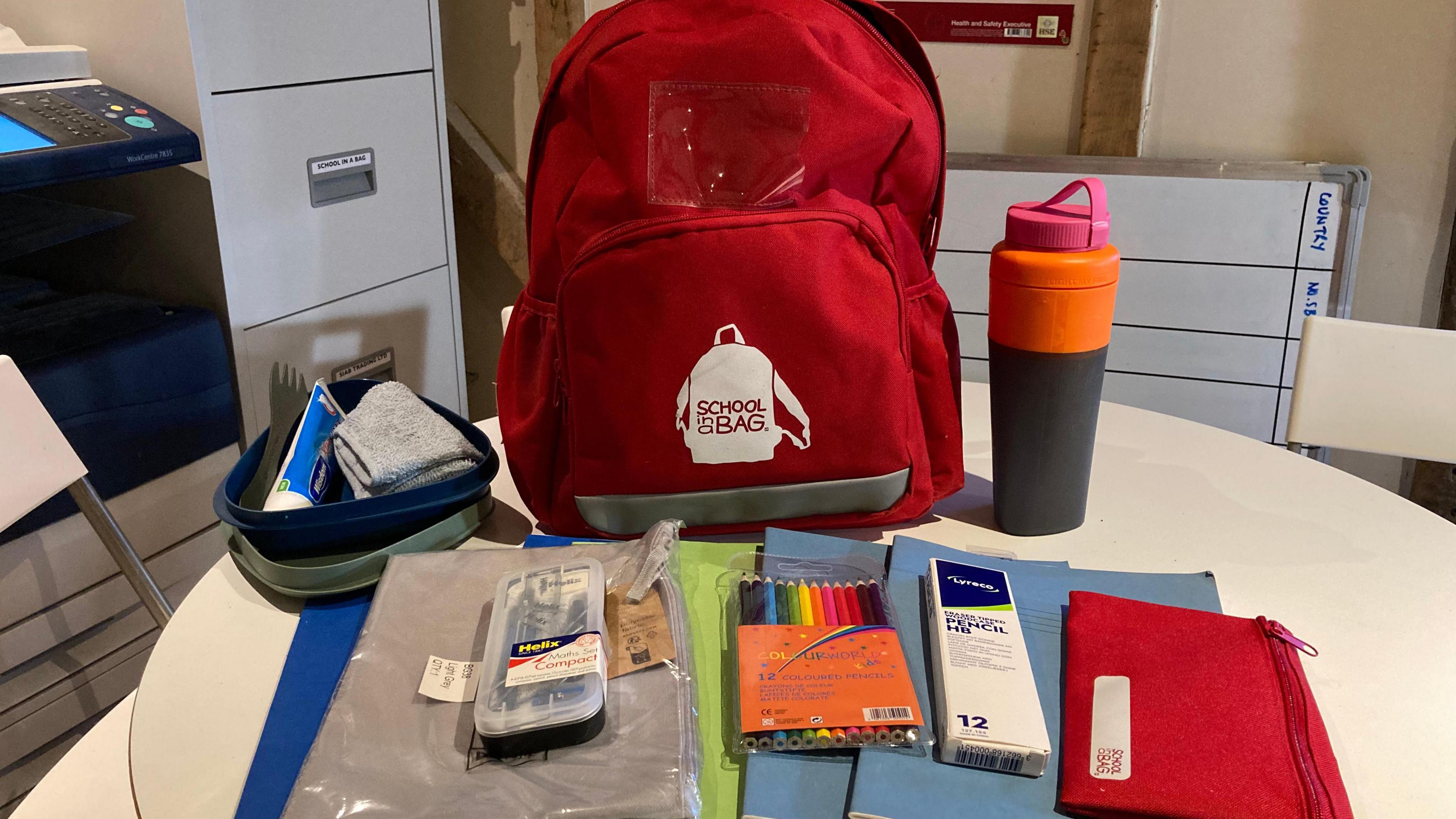 A red backpack sitting on a white round table. In front of it there are a selection of books and folders, a pencil case, washbag, school supplies and a water bottle.