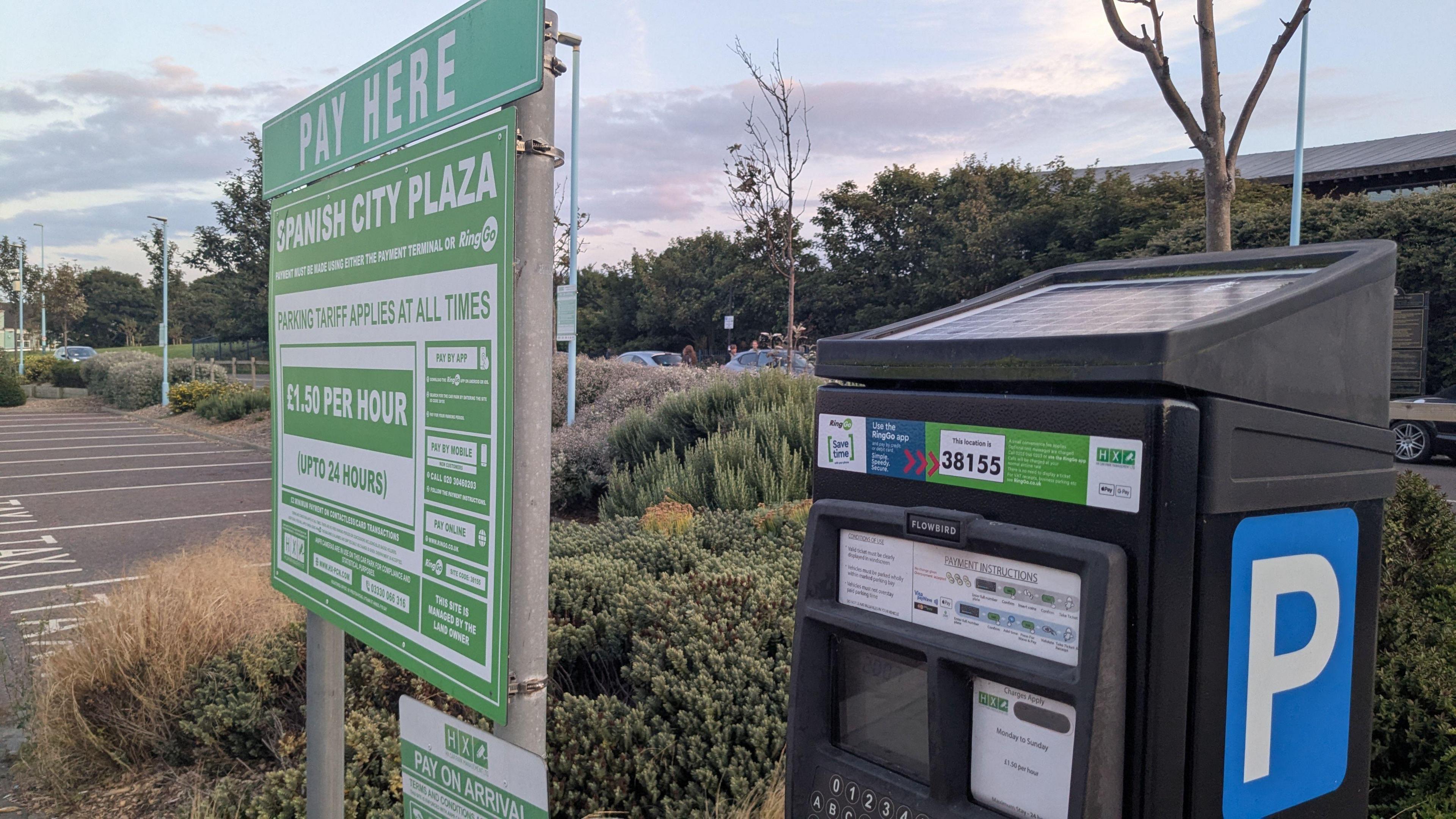 A black solar paneled pay and display machine stands in the foreground of a car park surrounded by tidily kept shrubbery. Next to it stands a green and white sign entitled 'pay here' with instructions and terms and conditions for parking on the site. 