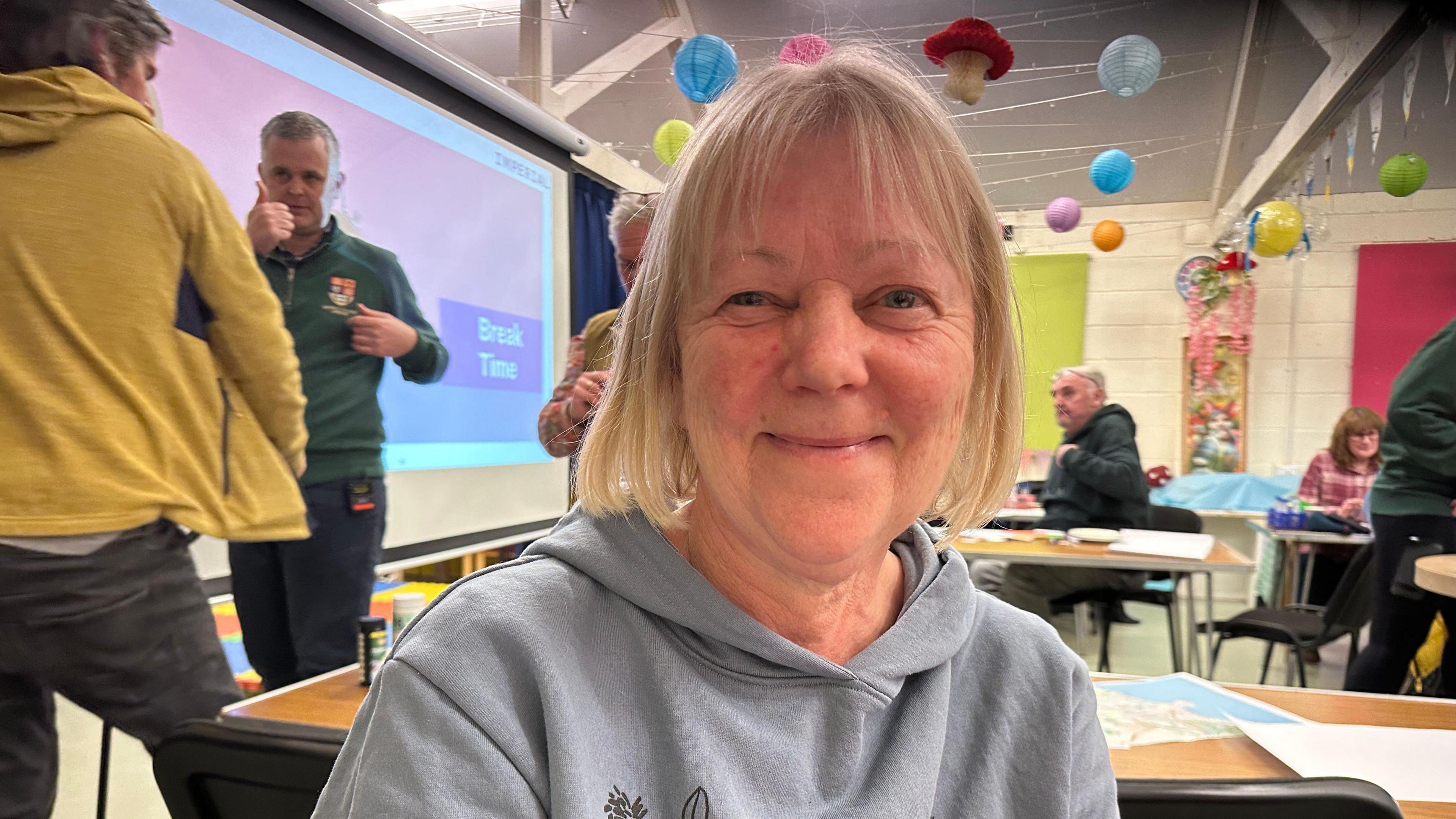 Woman with short blonde hair sits in a classroom and smiles at the camera.