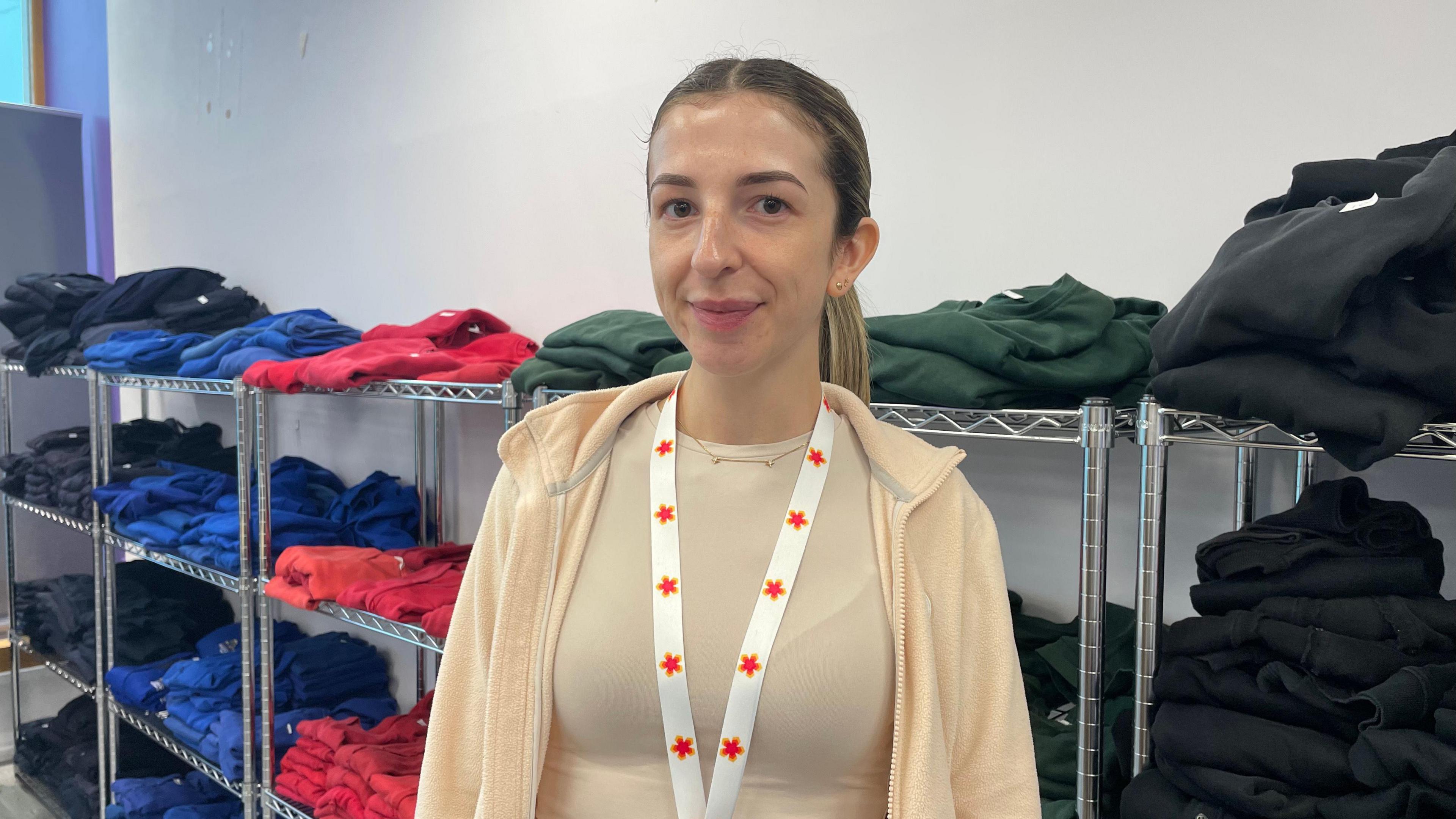 Paula wearing a beige hoodie and lanyard in front of a row of different coloured uniform.