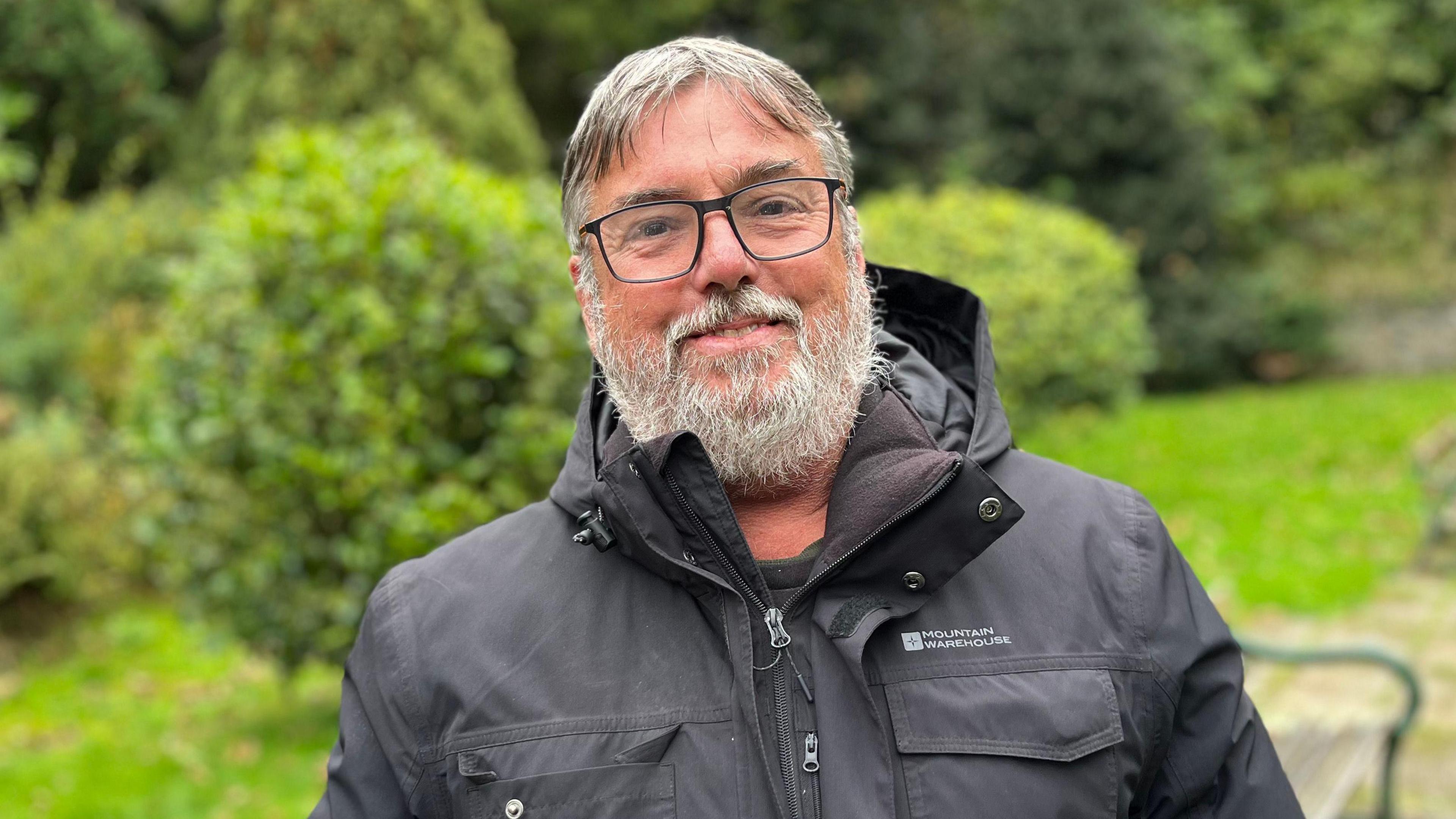 A man wearing a black coat, looking towards the camera wearing black glasses. He has grey hair with a middle parting and a grey beard. 