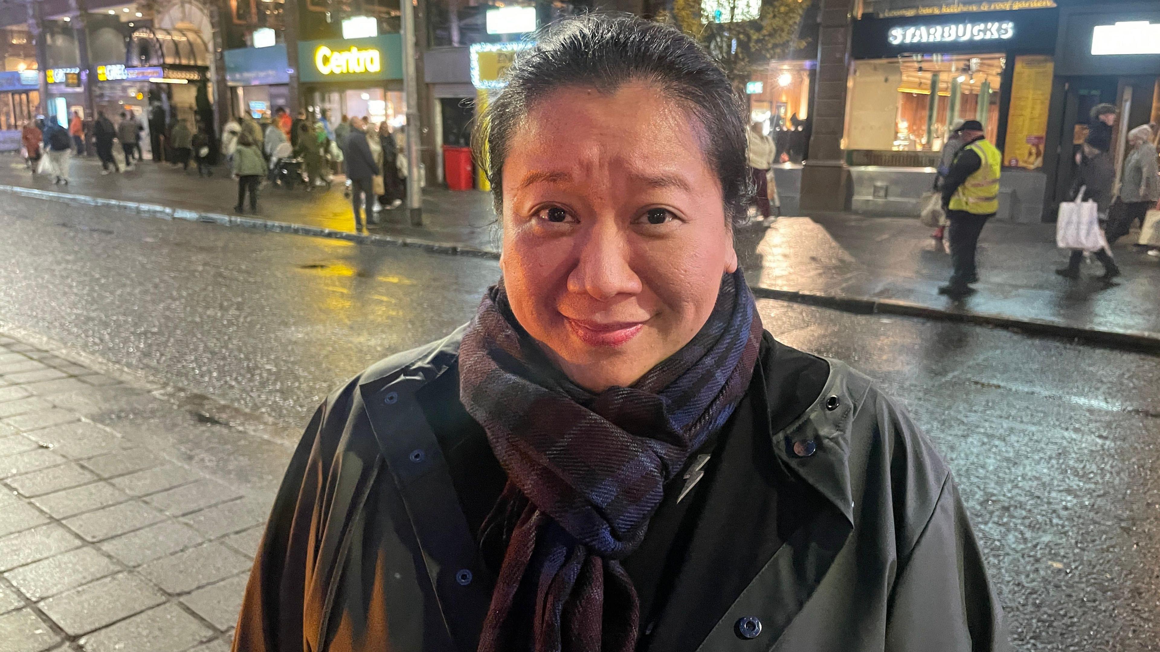 Tiffany wears a scarf and a dark coloured jacket. She has dark coloured hair tied up. 