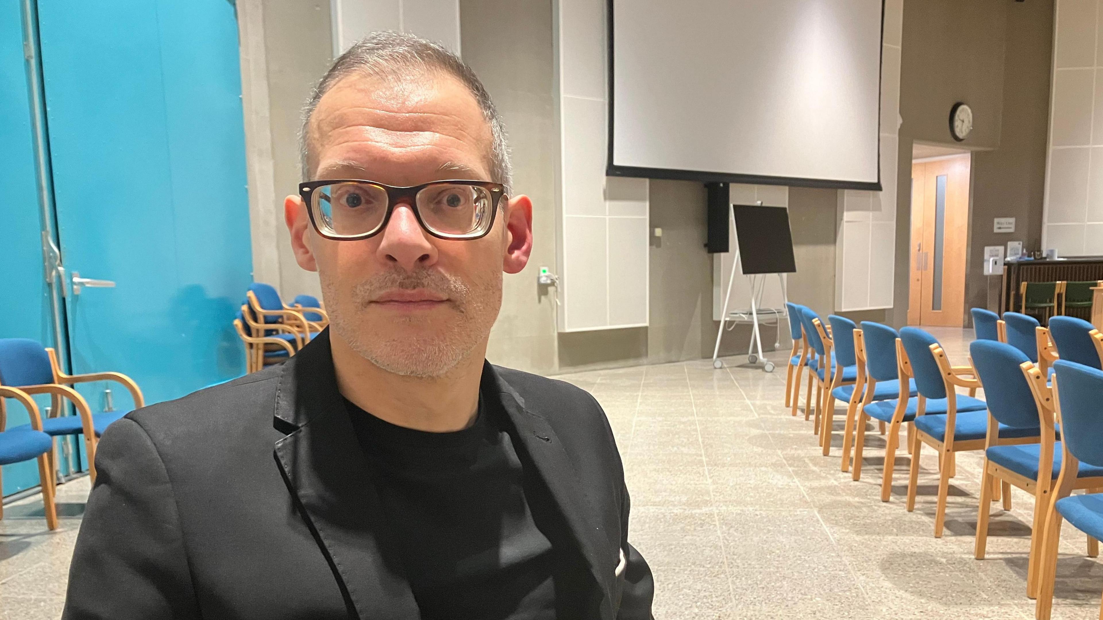 A picture of a middle aged man called Adam Zerny who is leader of Central Bedfordshire council. He is wearing glasses, has short hair, and has a black jacket on and a black t-shirt underneath.