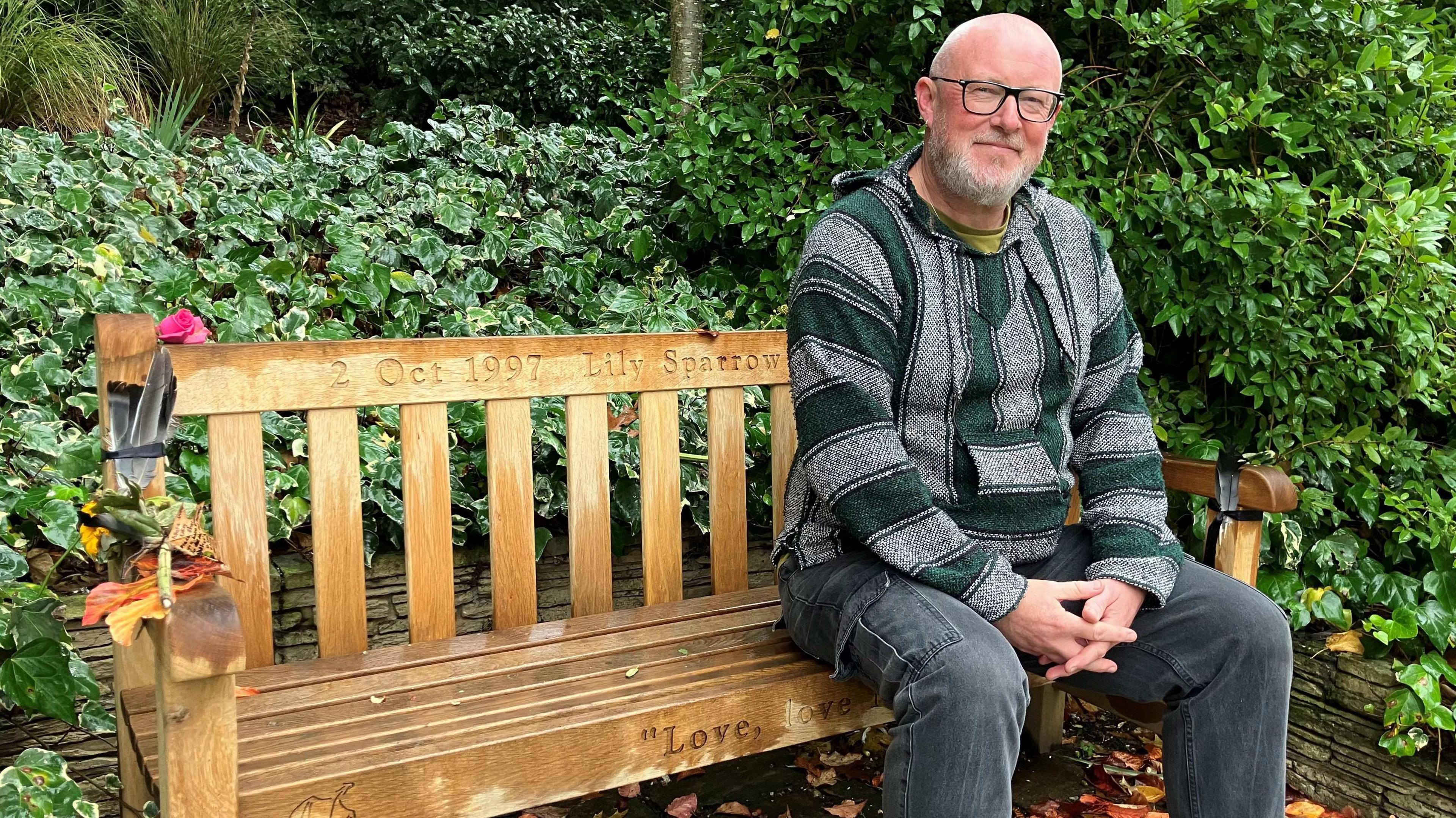 Simon Sparrow sits on a park bench, surrounded by trees and bushes. There are flowers and some feathers attached to the arm of the bench