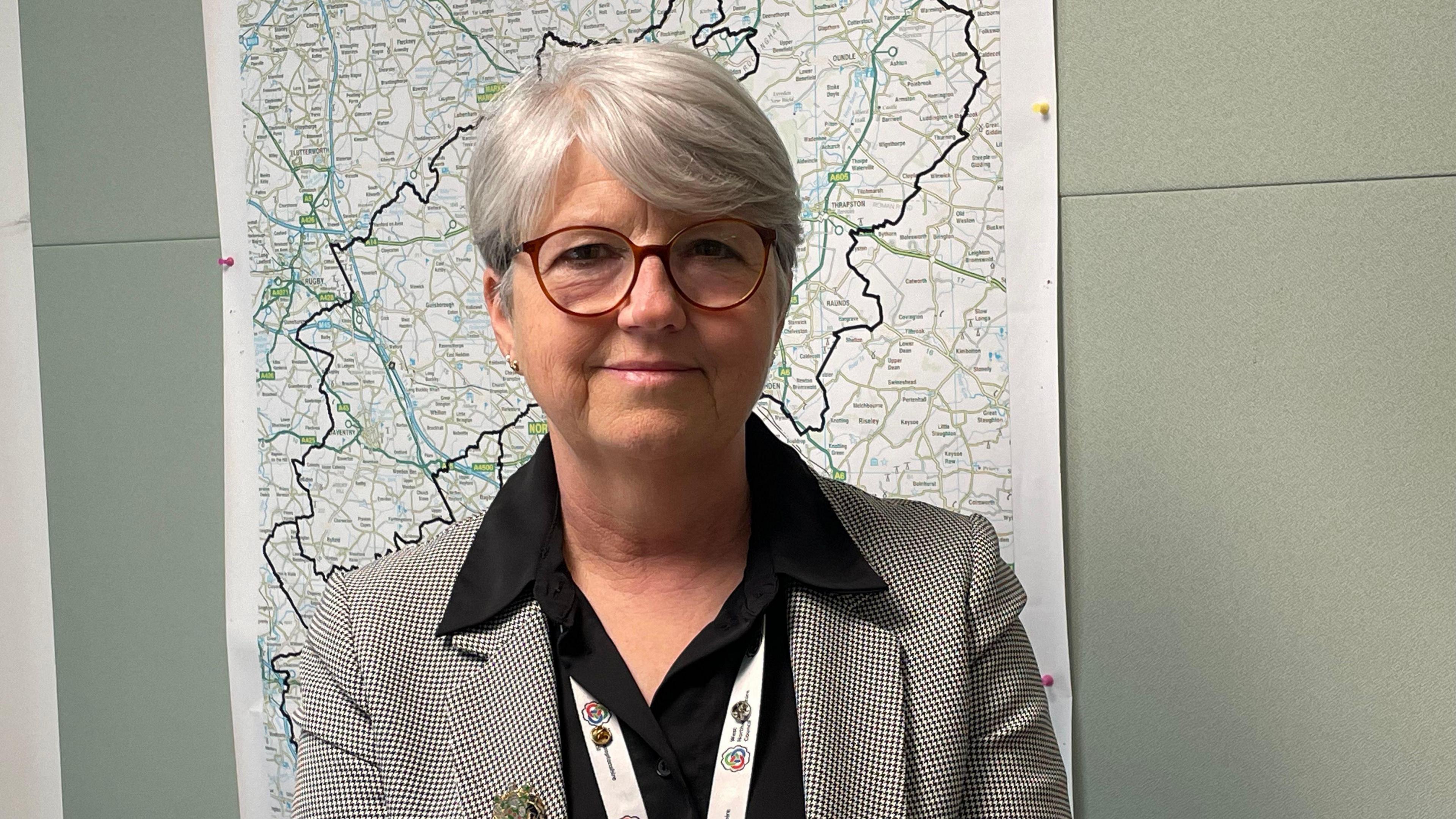 A woman wearing glasses and a back and white chequered blazer and black open shirt stands in front of a map of Northamptonshire. 