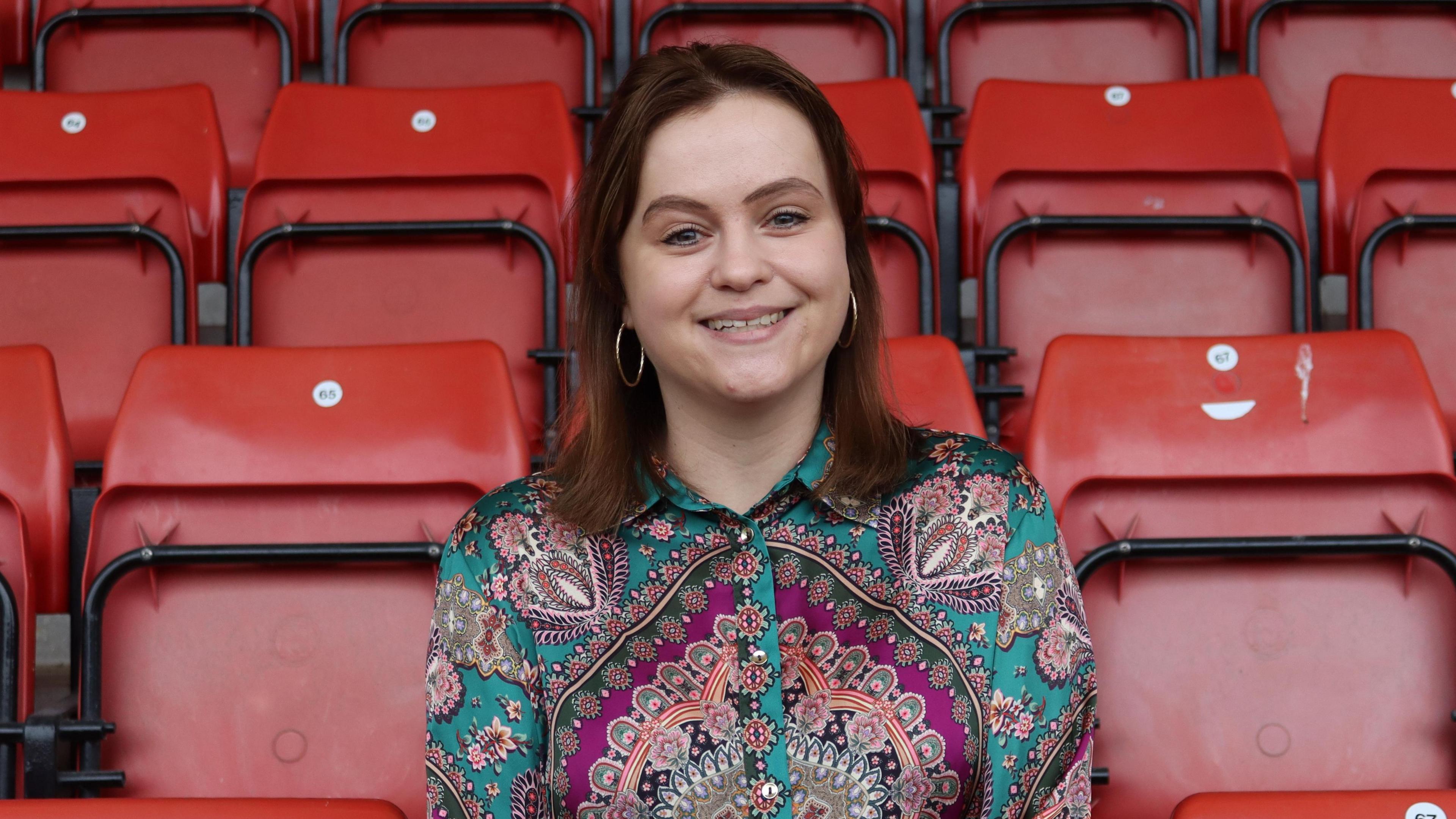A photo showing a woman with dark hair and wearing a colourful shirt. She is sat in a row of red seats in a football stand