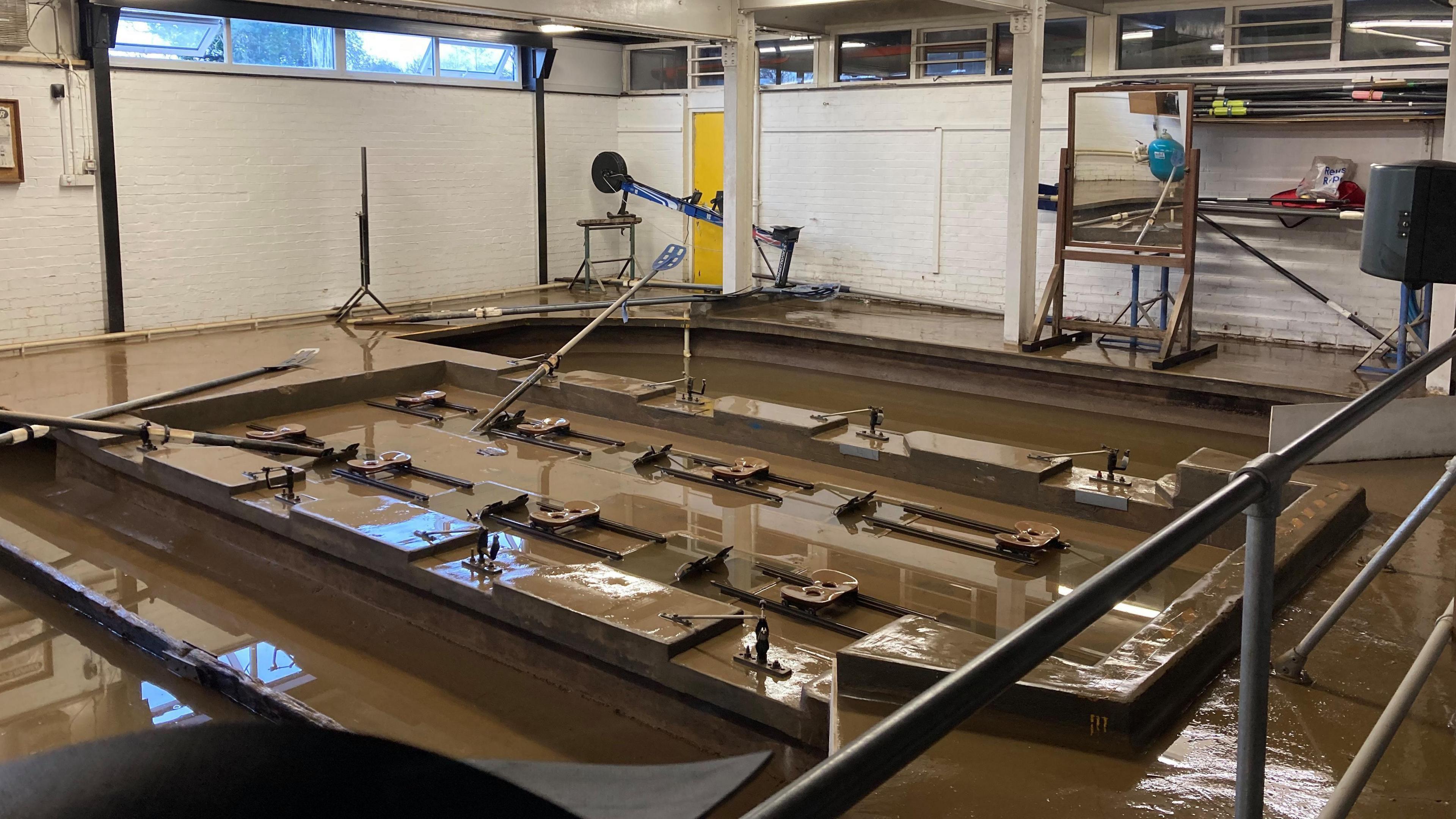 The inside of a room with several rowing machines, which are sitting in dirty brown flood water.