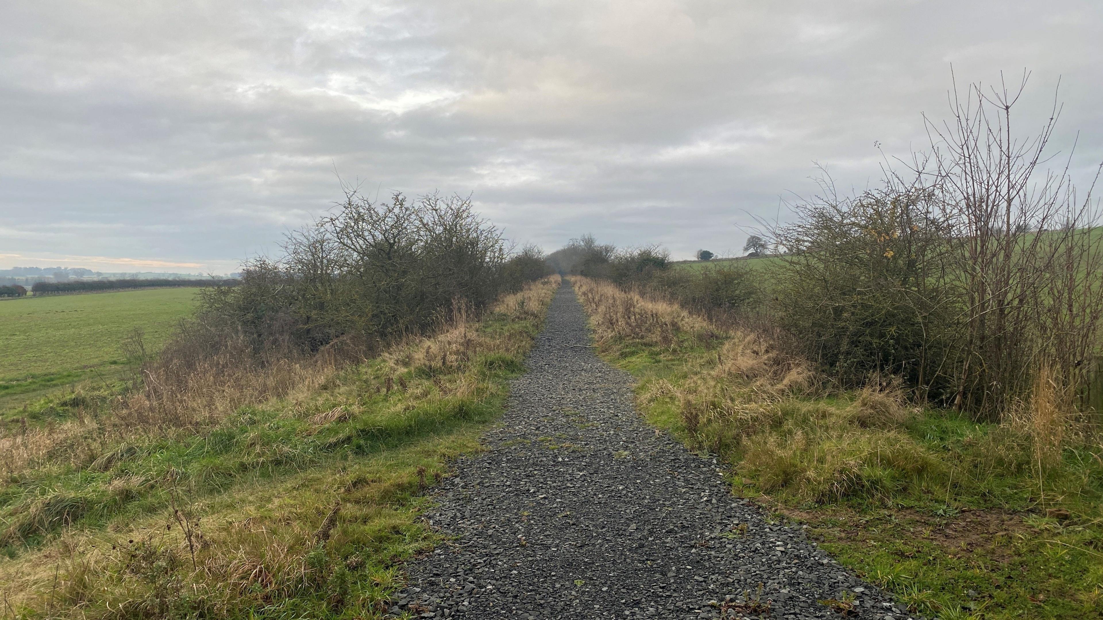 A former railway line has a restored path on it which is laid with stone. It crosses a flat area with fields on either side 