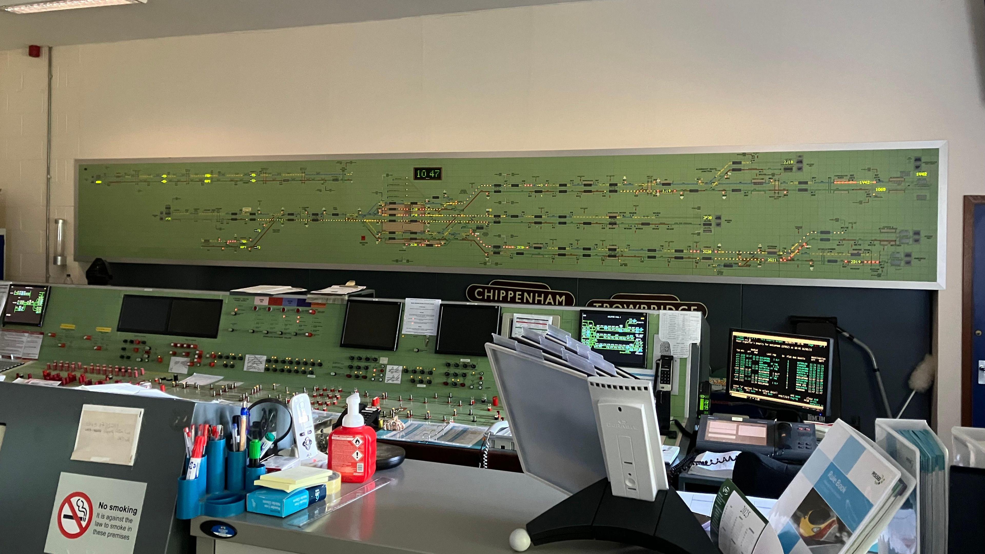 The inside of a signalling centre, which has a large green board on the wall showing the railway line and with various lights along it lit in green, orange and red. In front of it are computers and a control panel where the signaller works.