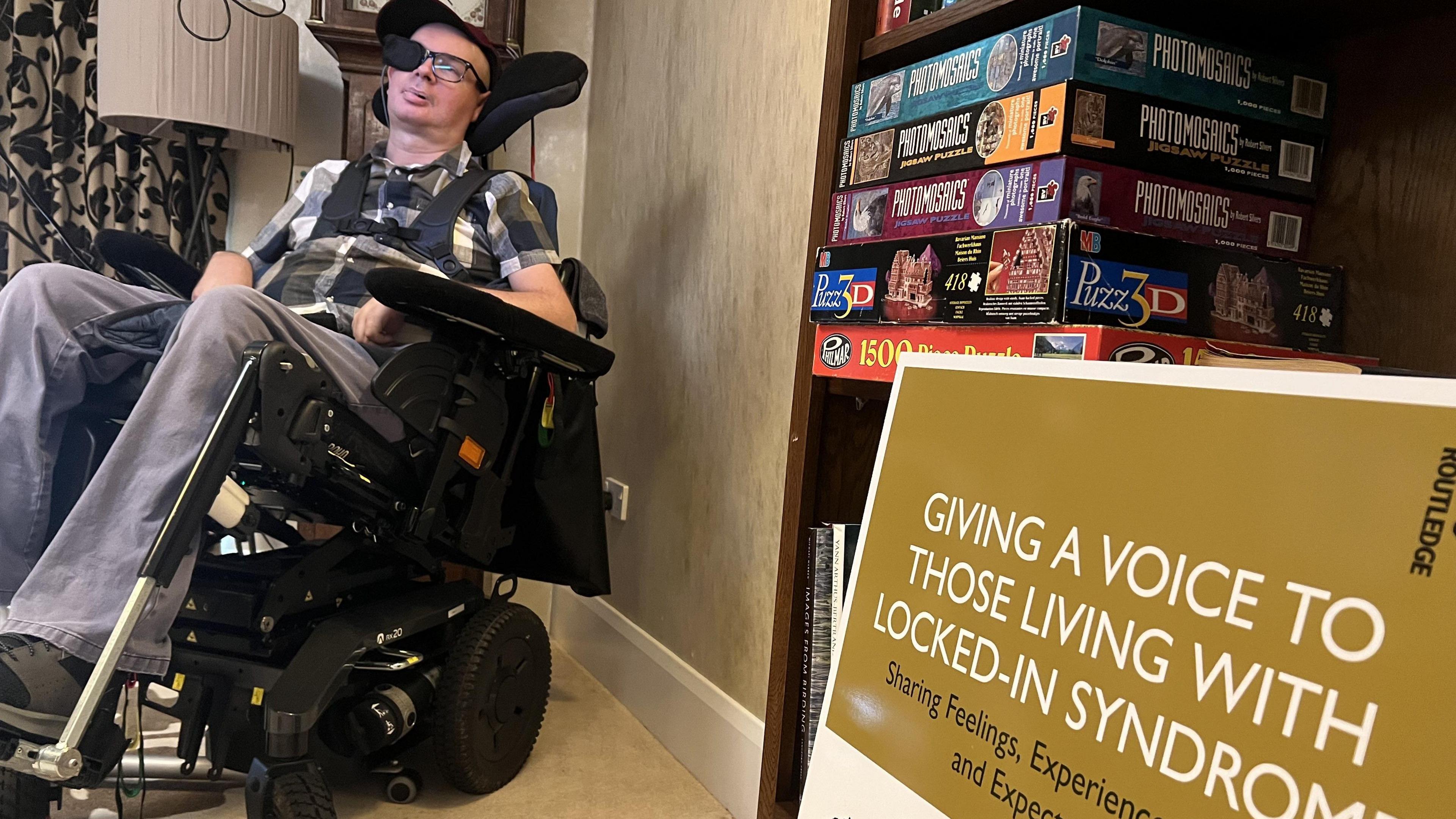 Bram Harrison in his wheelchair, in front of an eye controlled computer screen with a poster about about a book he has contributed to in the foreground.