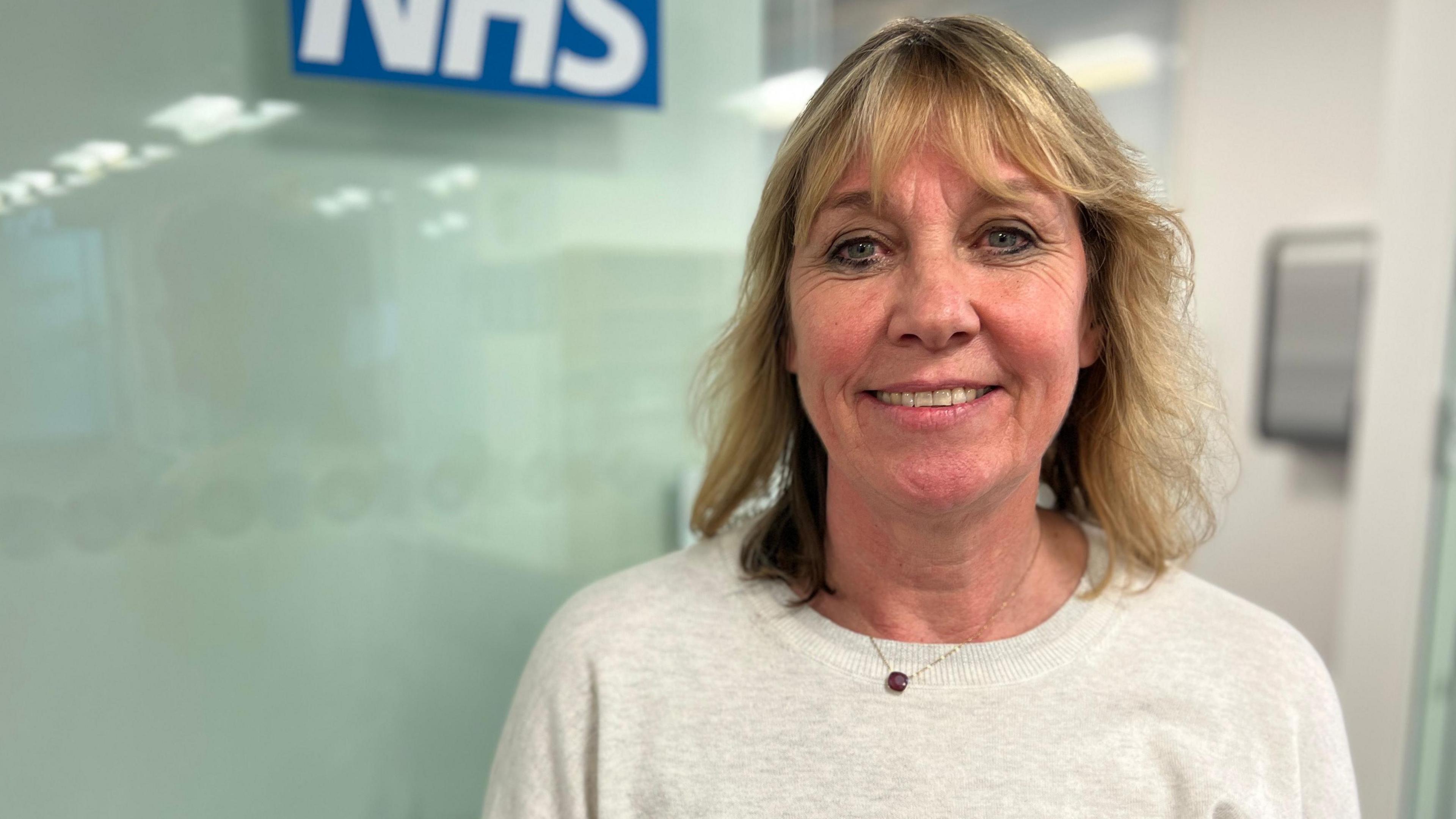 Gill May wearing a white blouse and small pendant necklace. She has shoulder-length blonde hair with a fringe and is smiling at the camera. She is standing in a hospital with a glass wall behind her that says NHS on it.