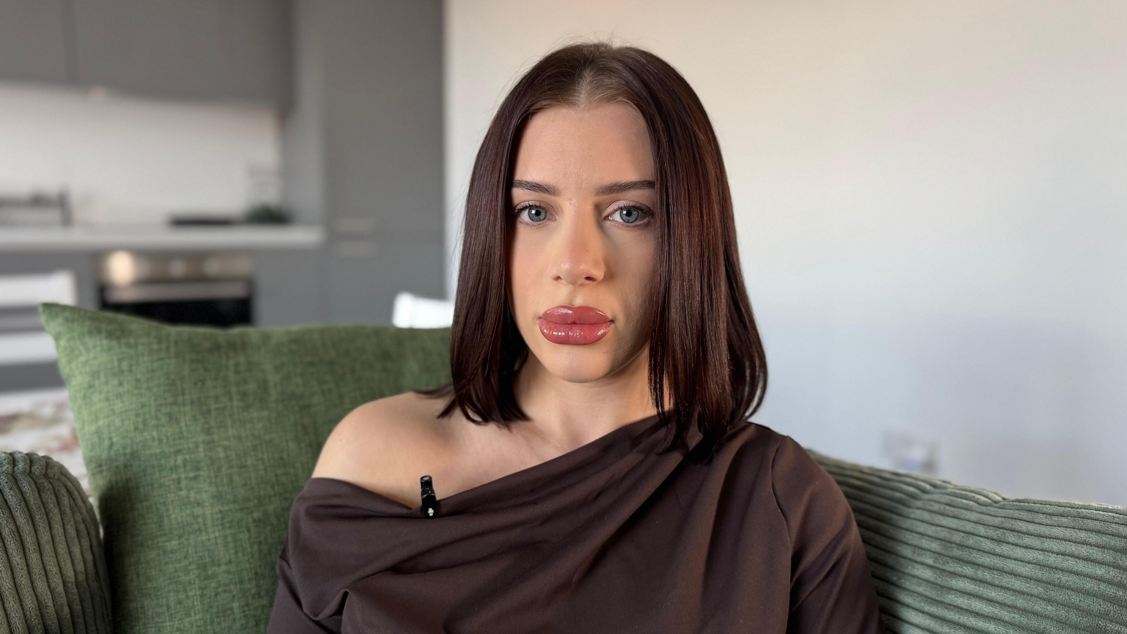 A woman sat on a green sofa. A blank wall and a kitchen can be seen in the background.