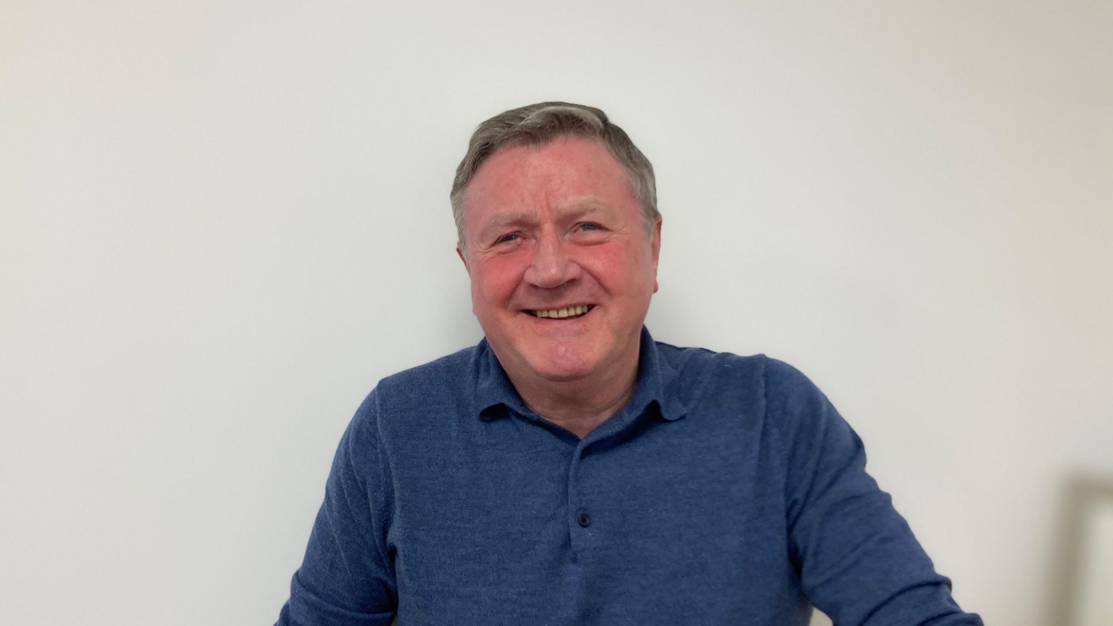 Dennis Jones stares at the camera with a smile on his face. He wears a navy top and stands against a white background.