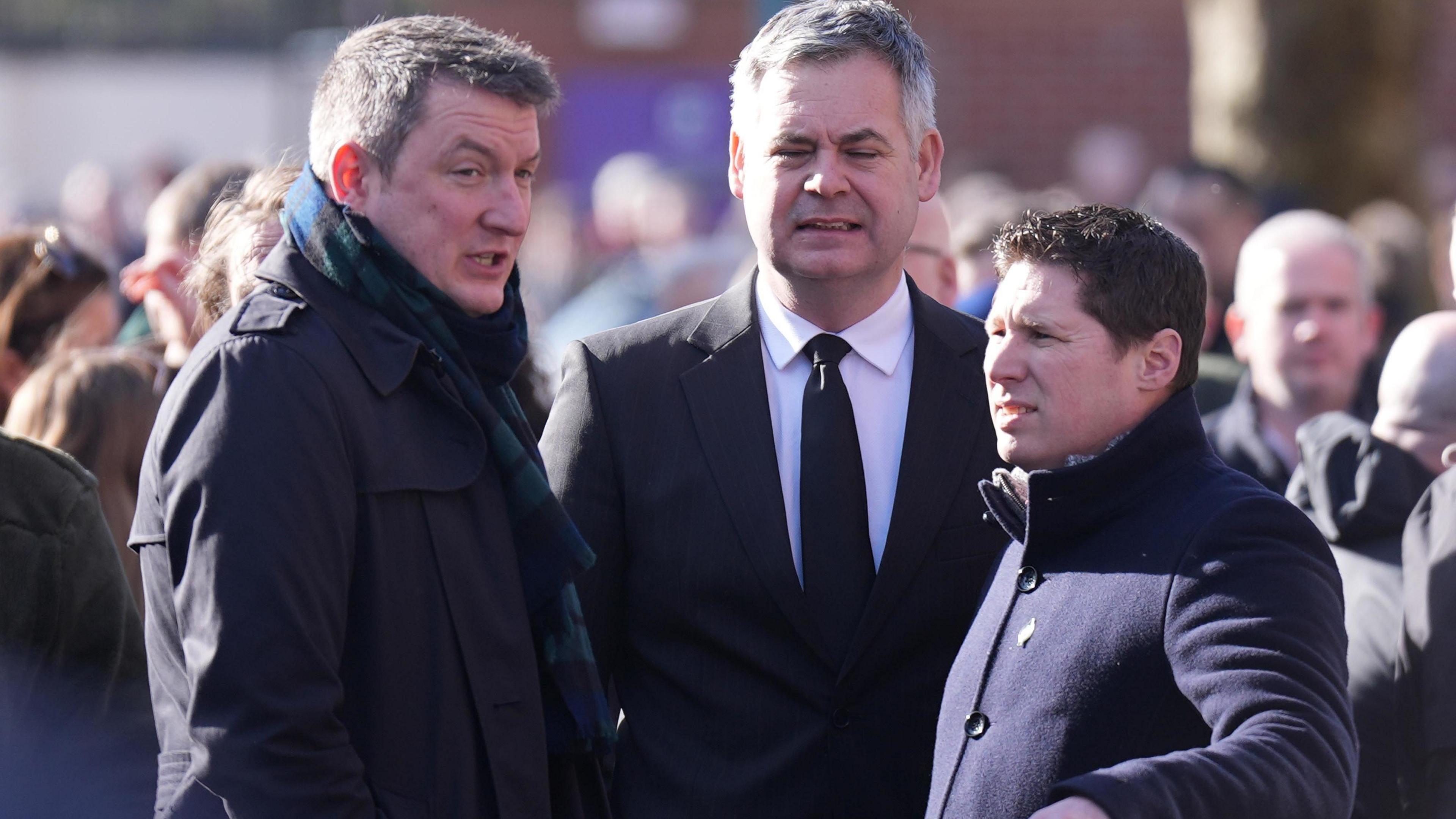 Three men among a crowd, the one to the left is wearing a black coat with a green, blue and black tartan scarf. He has dark grey hair. The man in the middle is wearing a black blazer with a black tie and white shirt. He has grey hair. The their man, who is at the right of the three, is wearing a navy coat. He has black hair.