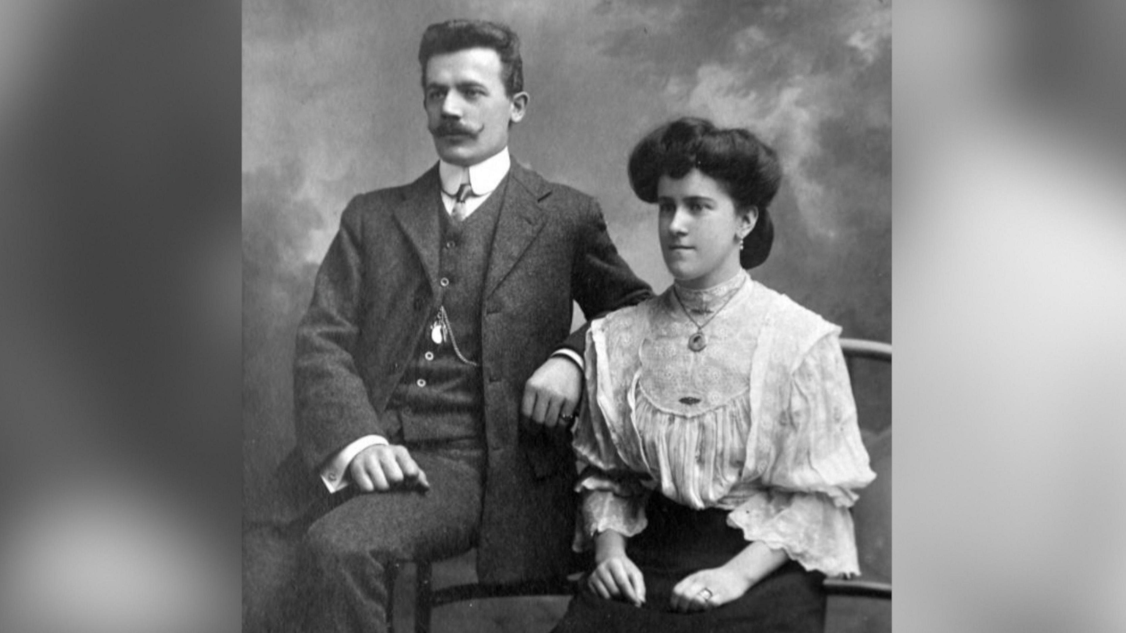 Max and Mabel Wulff, a Welsh-German couple, are photographed in black and white. Sat side by side Max and Mabel are dressed smartly in a three piece suit and a white blouse with a skirt respectively. 