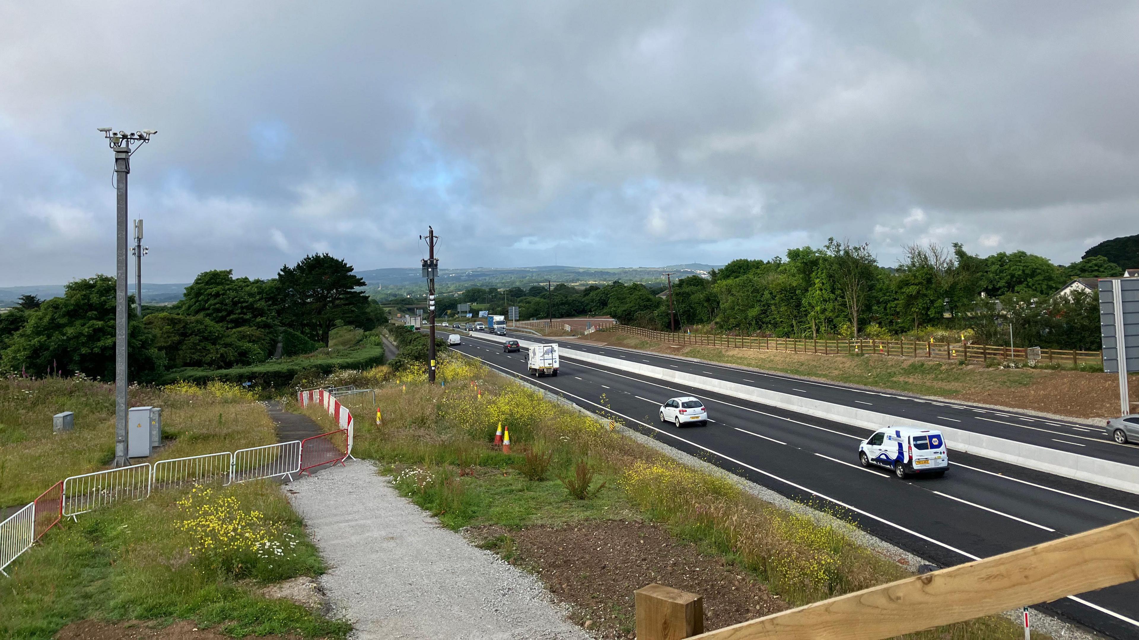Cars and vans travel along the A30 in Cornwall on a cloudy day. Grass verges and fields are on either side of the dual carriageway.