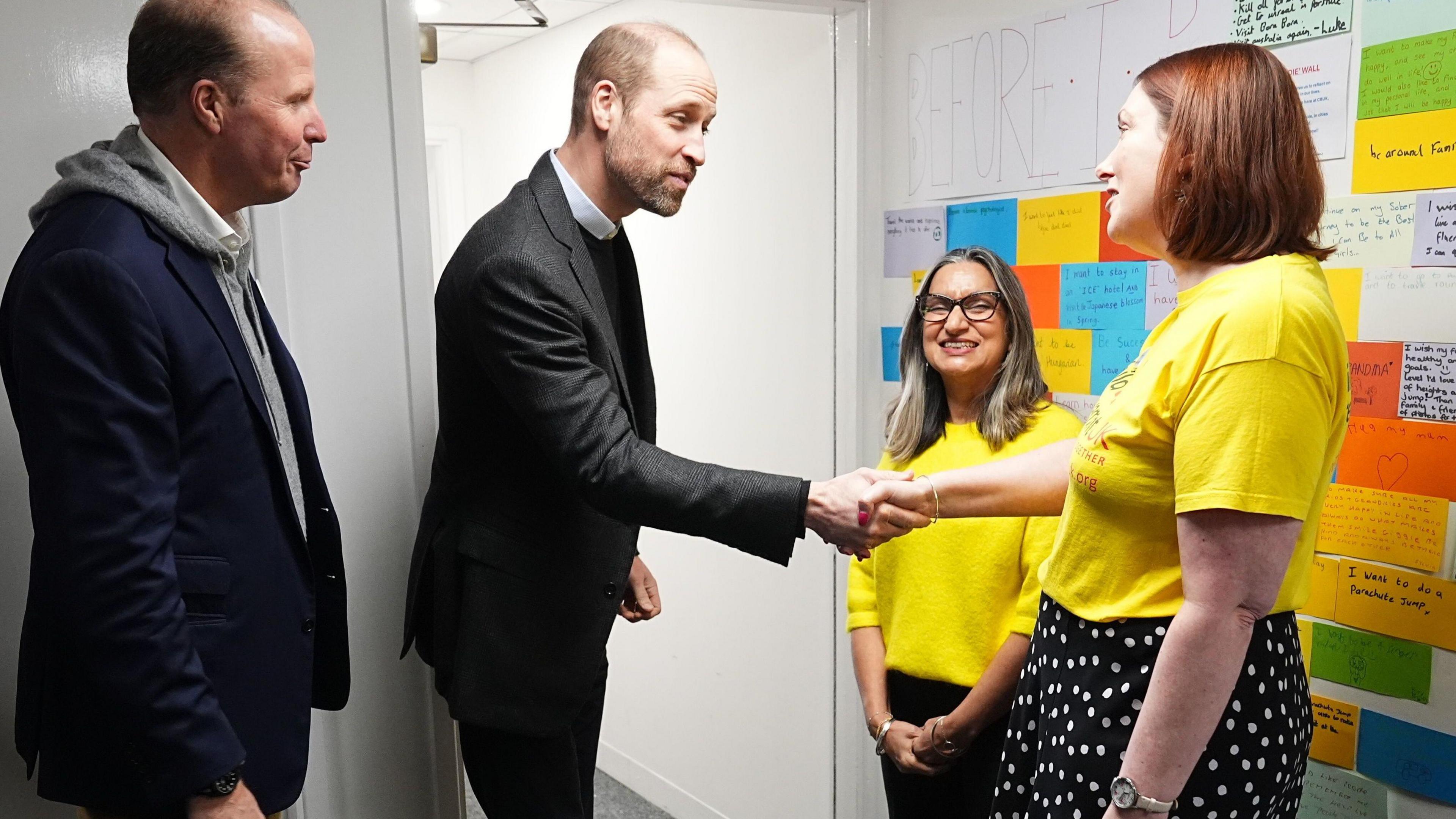 Prince William meets two women from the charity. He shakes hands with a woman wearing a yellow charity t-shirt. She is wearing a black polka dot skirt.