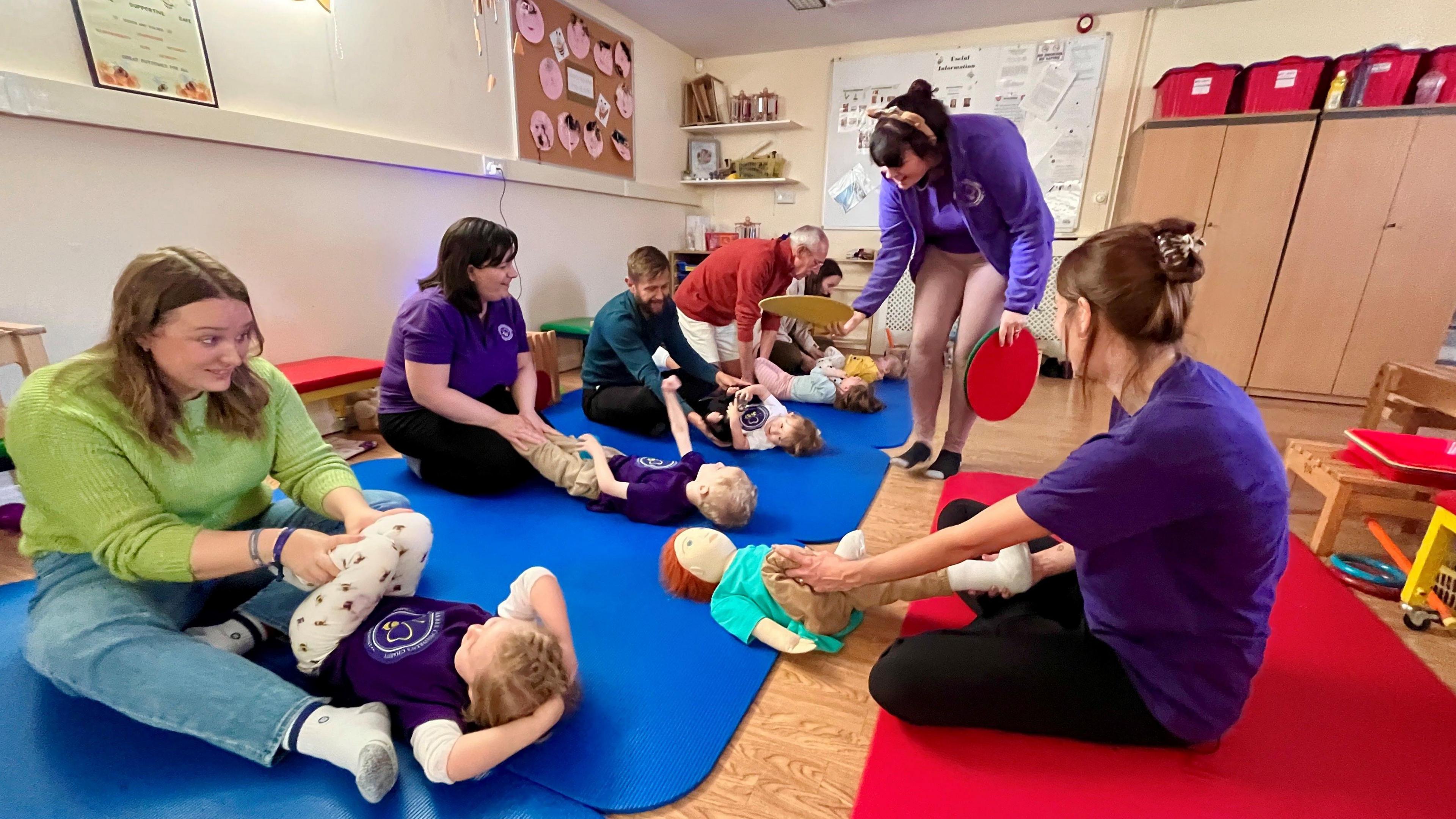 Adults, including some in branded purple tops, are doing motor exercises with young children who are lying on their backs on mats.