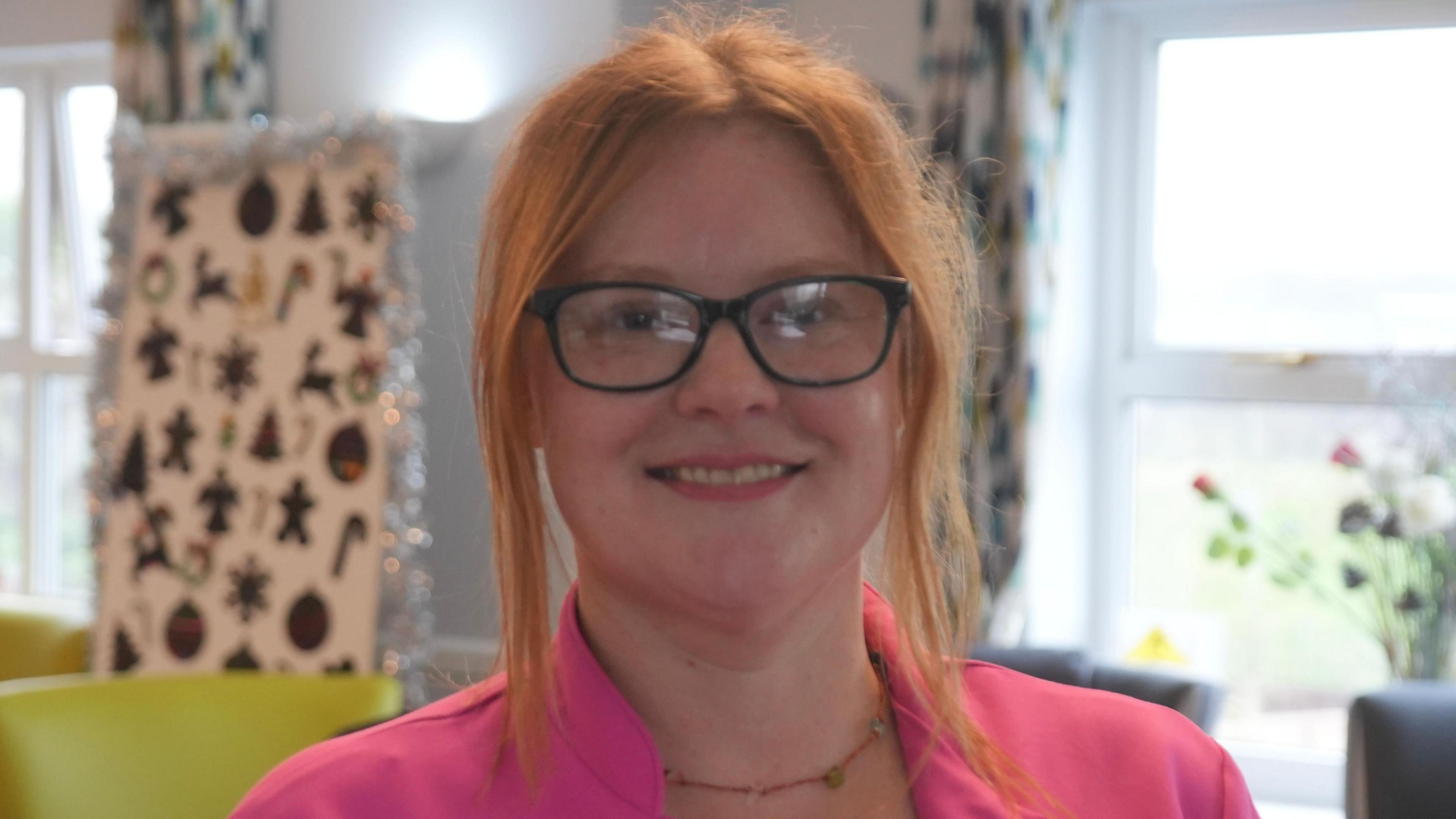 Alex Kelly, who is wearing a pink top while standing inside a room at the care home. She has ginger hair and is wearing glasses while smiling.