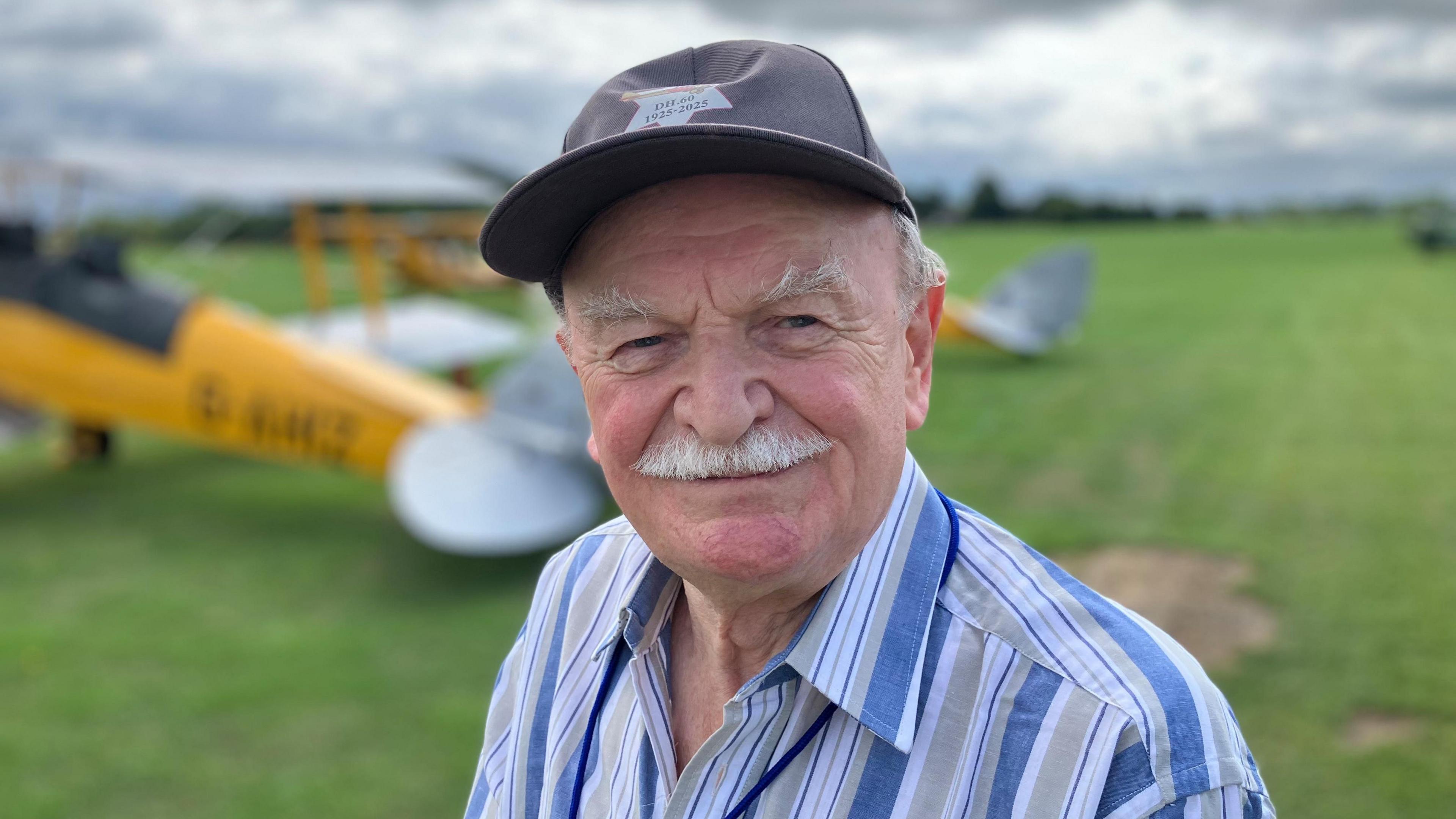 An older man in a striped shirt and dark hat standing on a grassy field with a yellow biplane behind him.