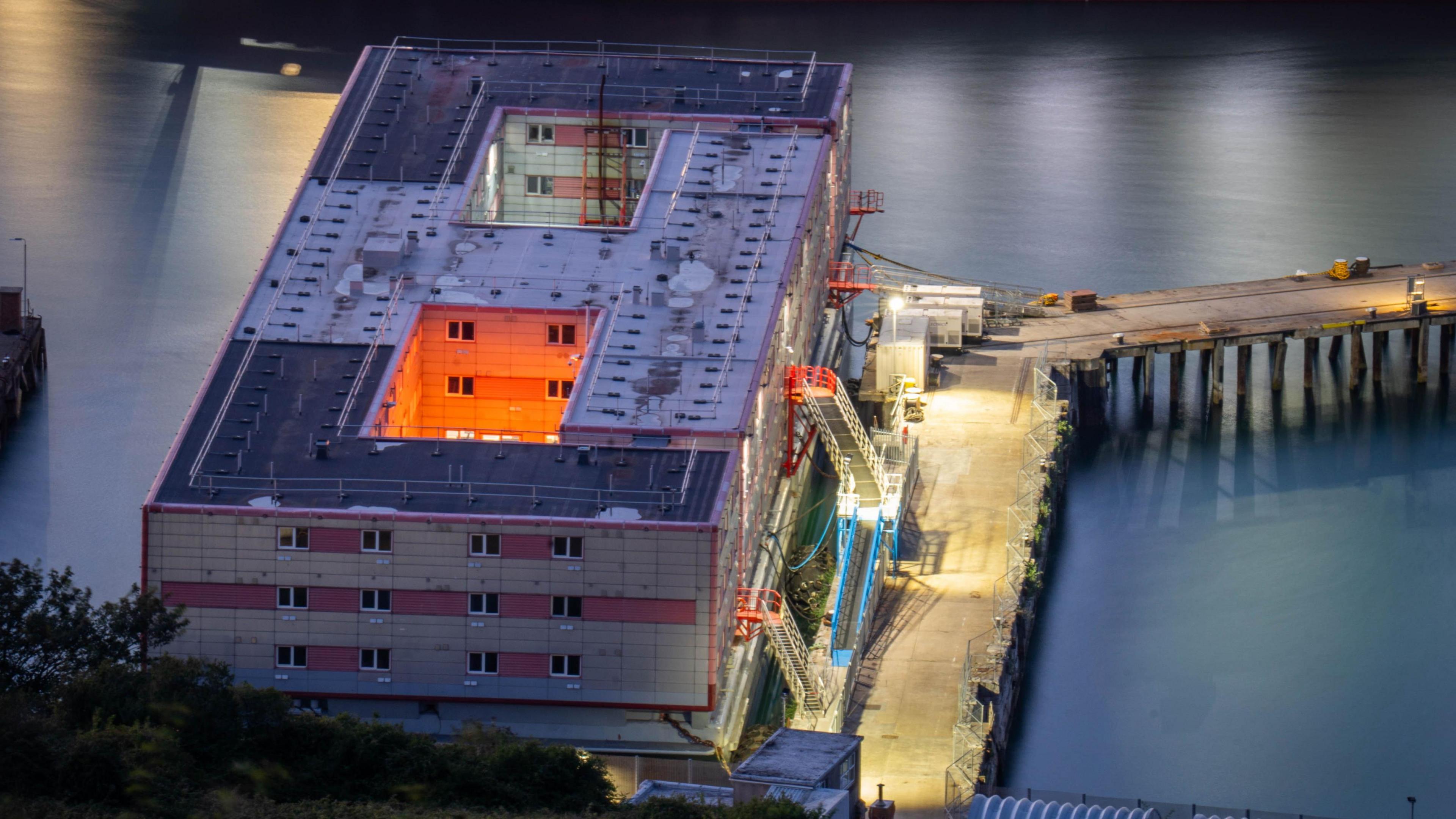 The grey, rectangular shape of the Bibby Stockholm at twilight. Orange lights illuminate the interior courtyard of the vessel, while white lights highlight the dockside and water.