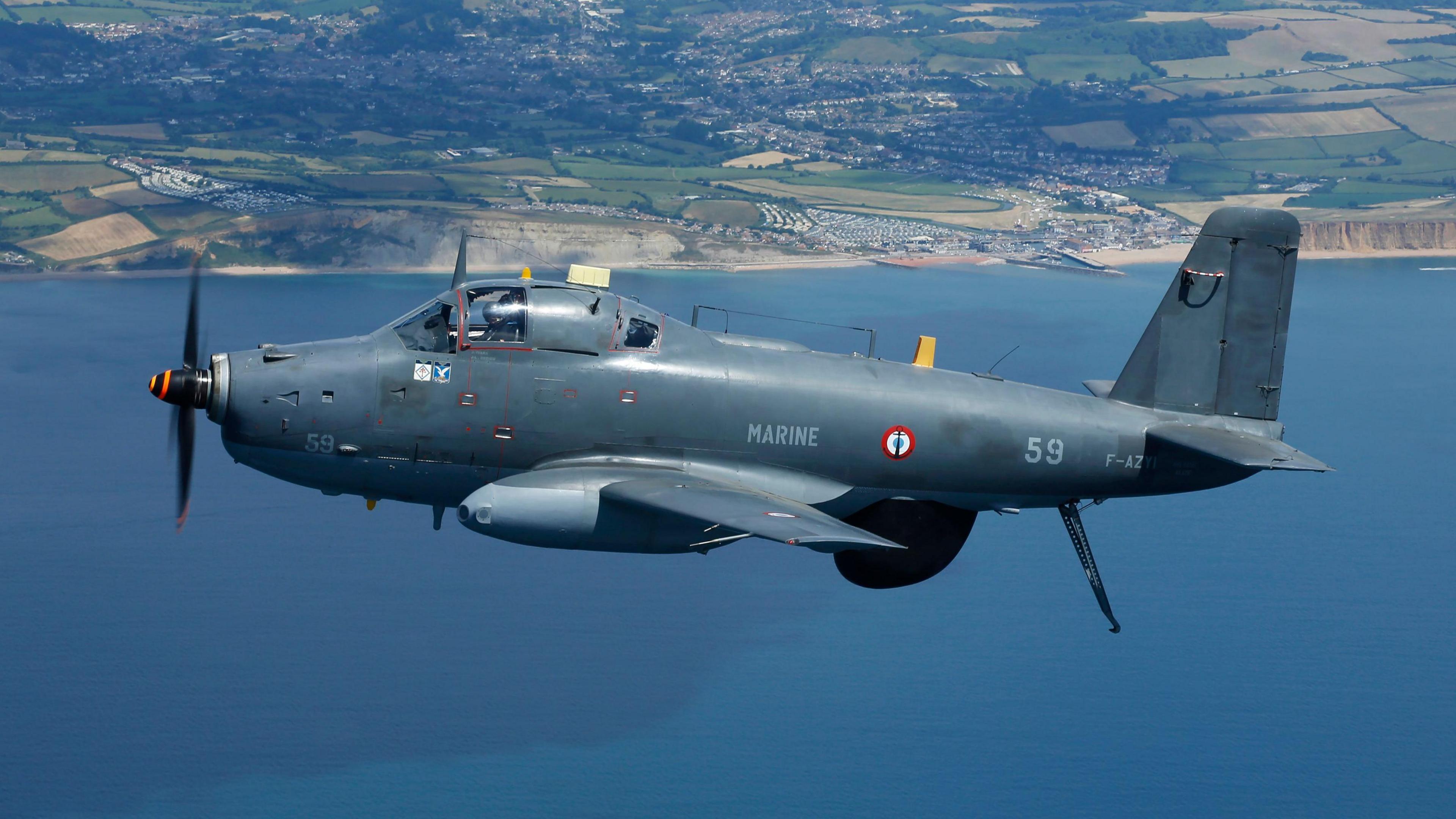 A French Breguet Alize anti-submarine aircraft flying above the sea with a coastline in the distance. The grey plane has the word MARINE printed on it side.