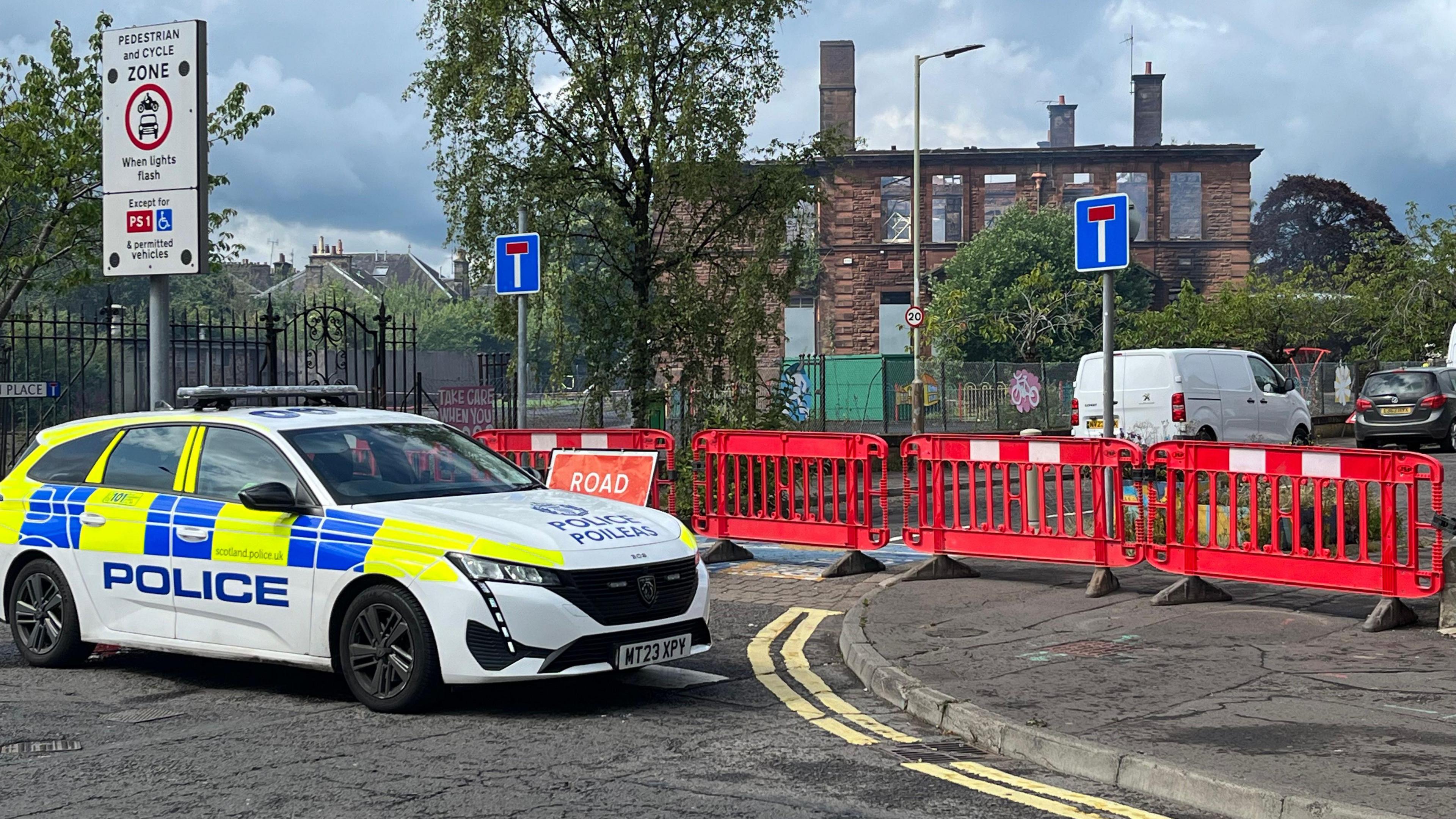 A police car parked in front of the school. The roof has been worn away by the fire. There is a red plastic fence in the foreground, parked behind a police patrol car.