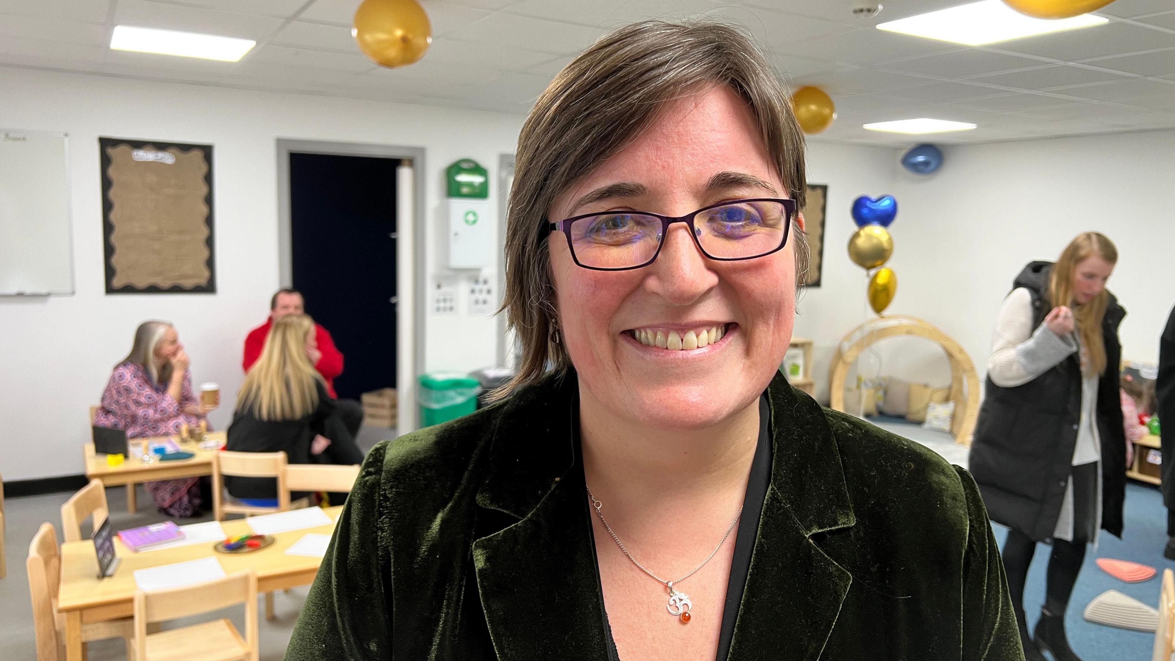 Catherine McLeod, who runs the charity Dingley's Promise, is wearing glasses and a green jacket. She is smiling at the camera. The photograph has been taken inside one of the centre's play rooms so there are small wooden chairs and tables behind her along with lots of children's toys.