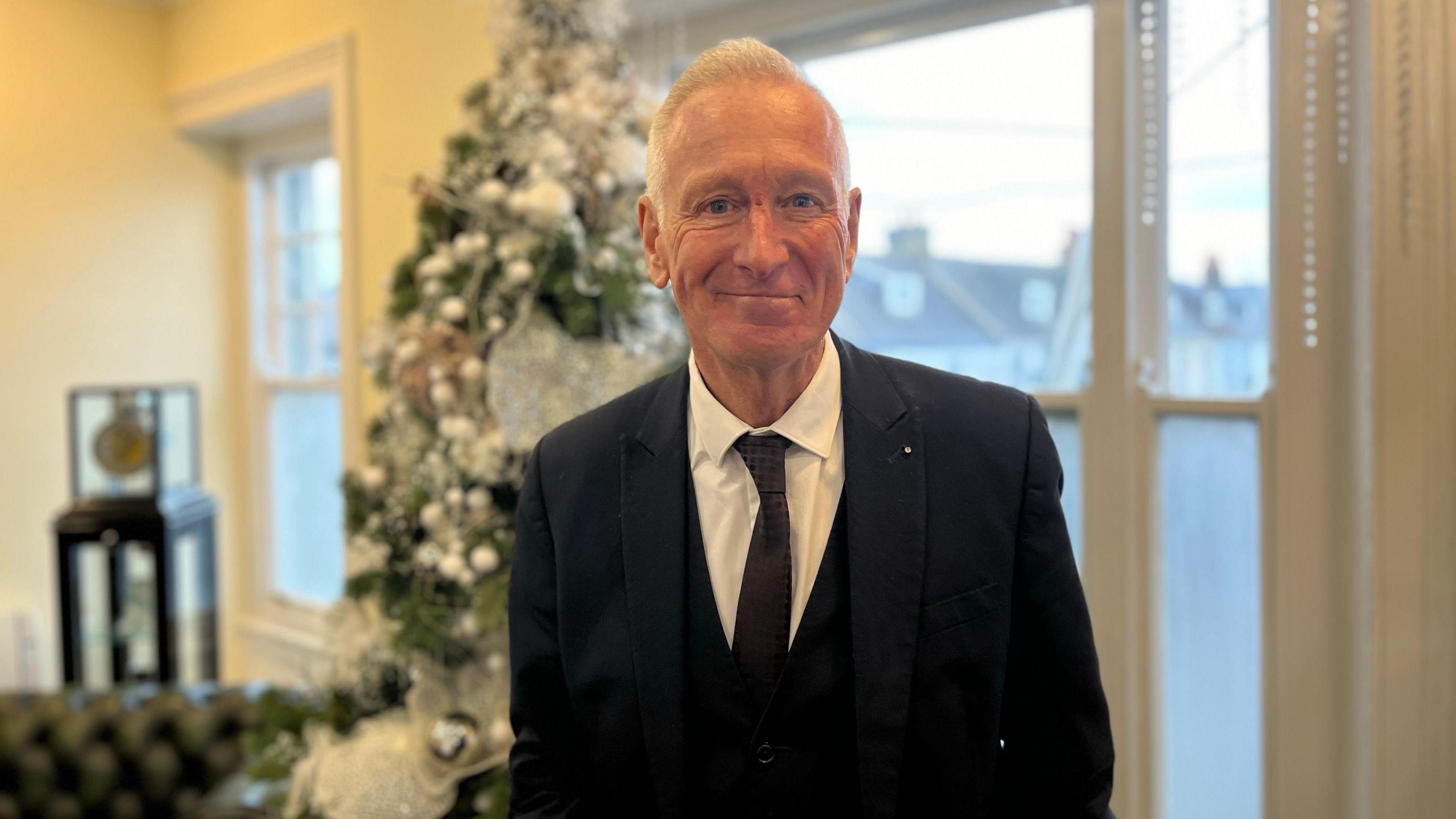 Martin Search dressed in a navy suit, waistcoat, white shirt and blue patterned tie. He is smiling without teeth and looking at the camera. He has white hair and there is a silver decorated Christmas tree in the background along with a window. 