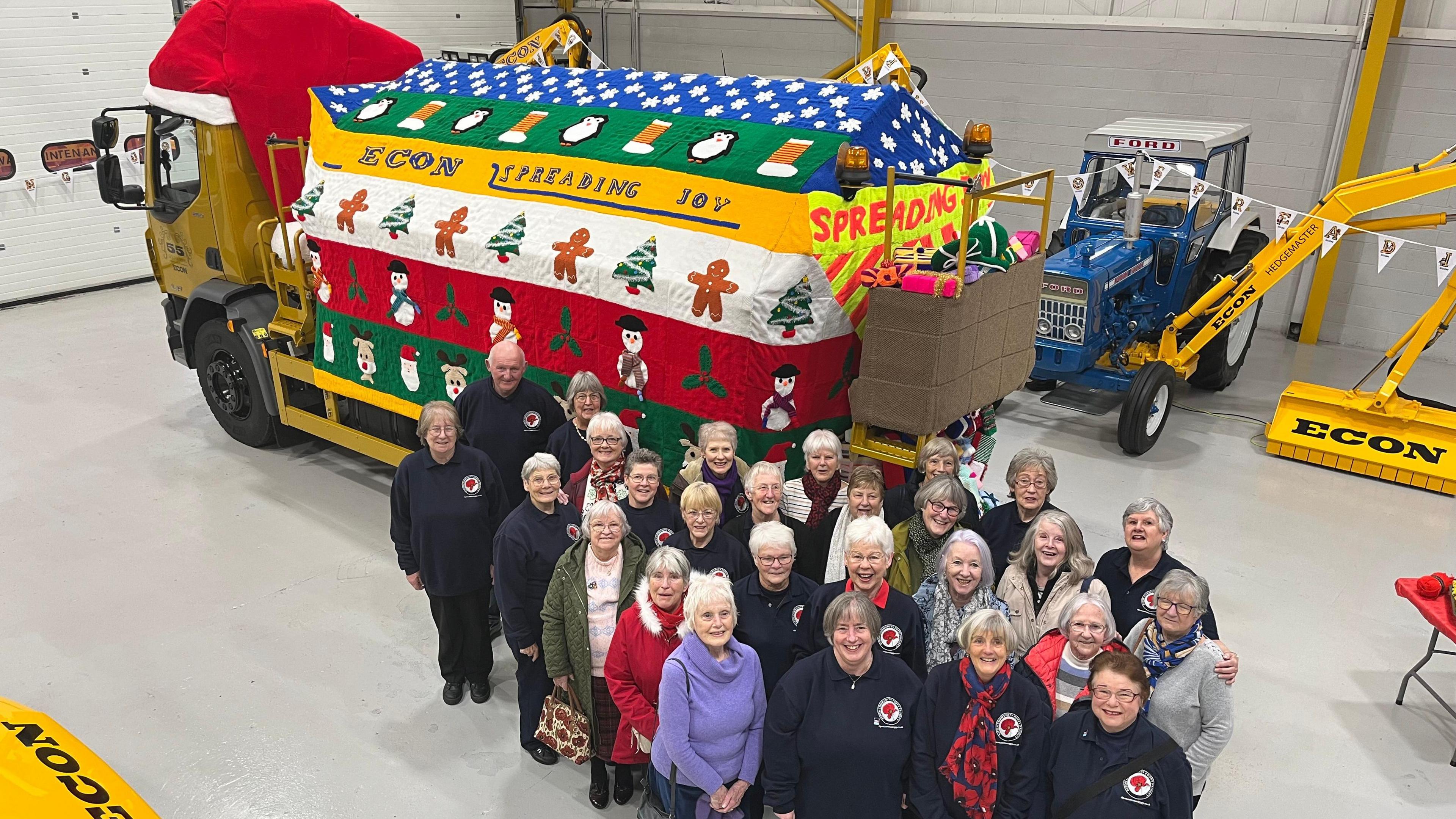 A group of knitters, standing in front of the gritter covered in a Christmas jumper, look up at the camera. 